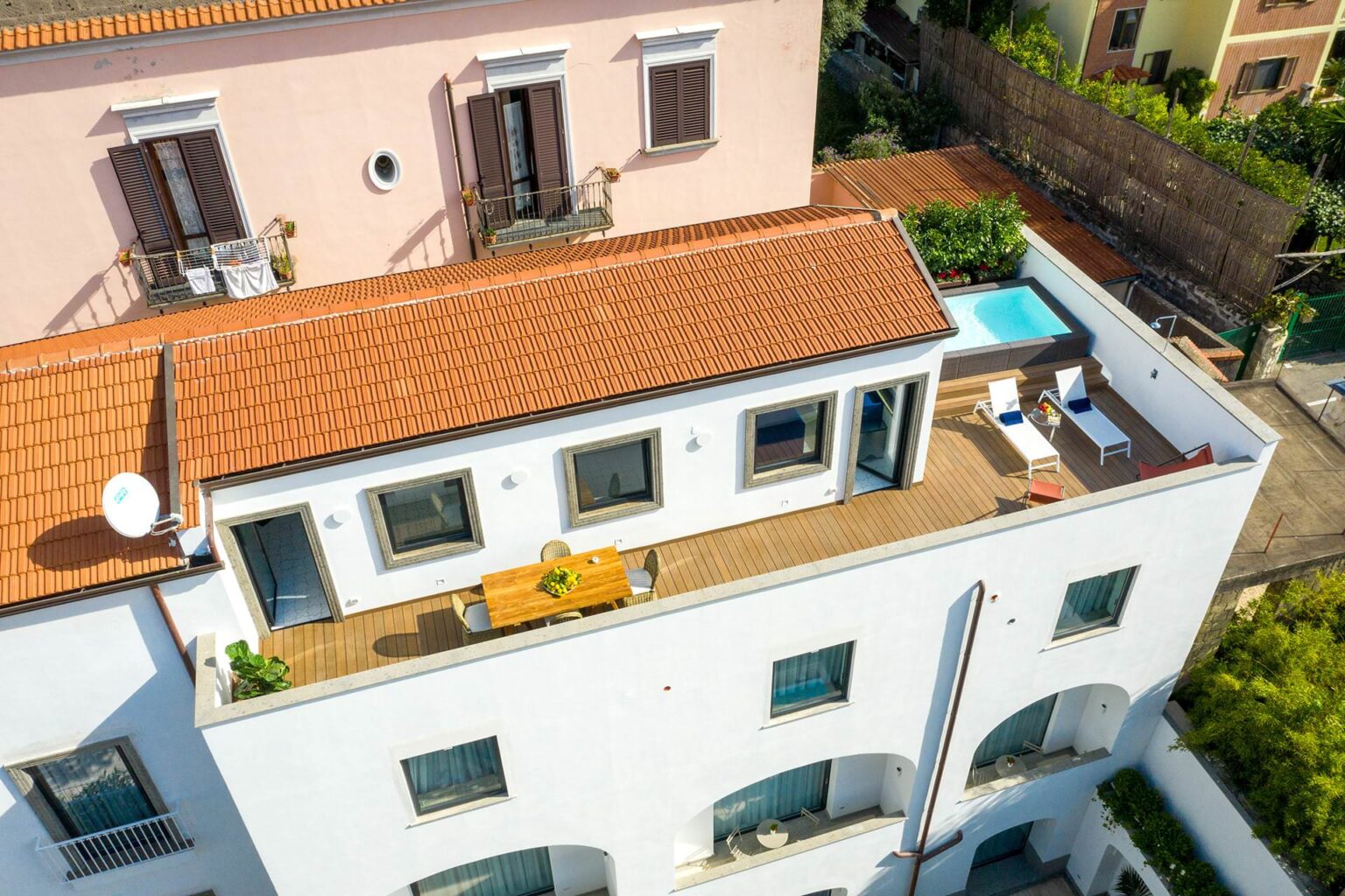 Aerial view of a modern white villa with a terracotta roof, featuring a spacious terrace with outdoor furniture and a small swimming pool. Lush greenery surrounds the property, enhancing its appeal in a scenic location. | Only on SopranoVillas