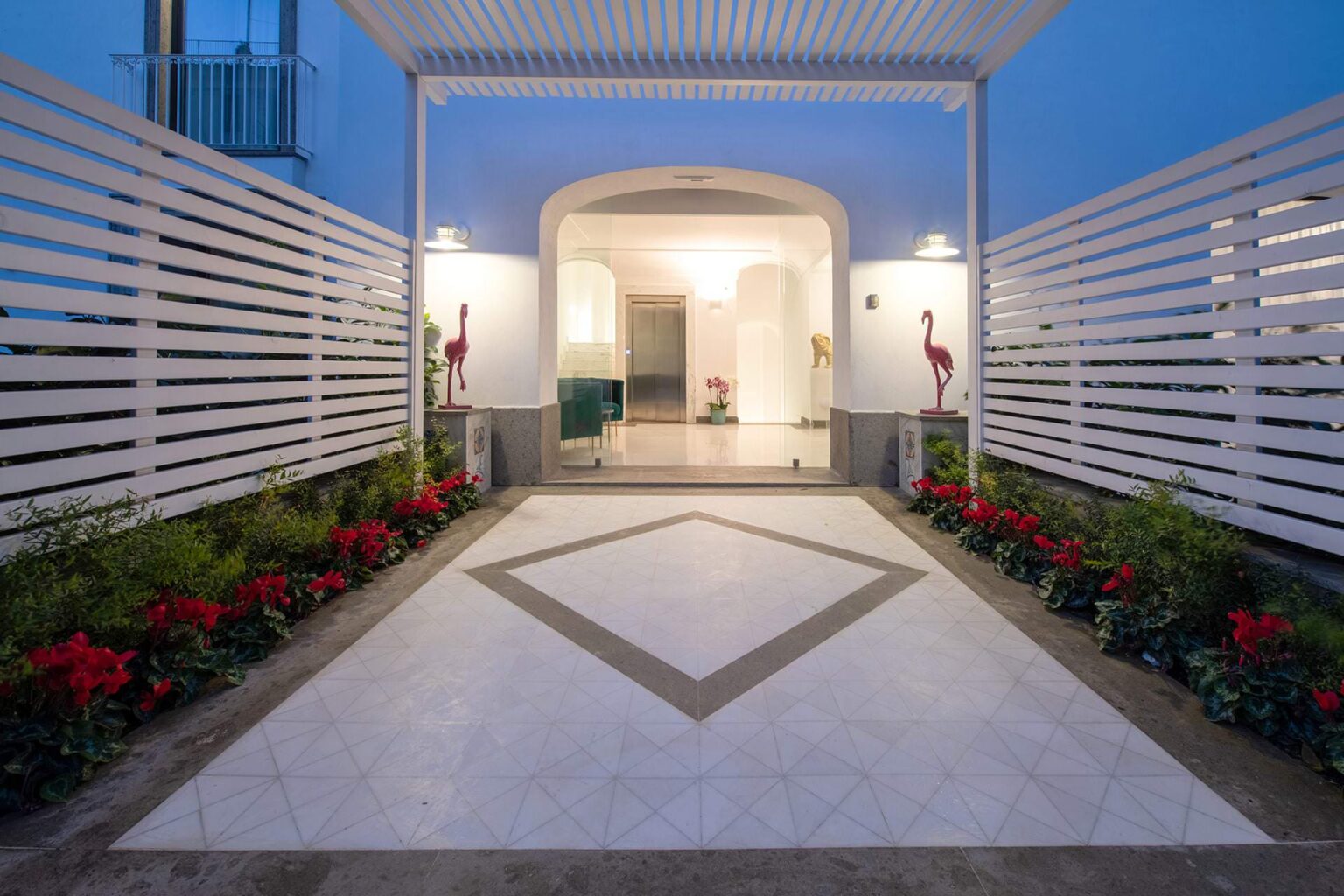 Elegant entrance with a geometric tile design, surrounded by vibrant red flowers and decorative flamingo accents, leading into a well-lit interior space. | Only on SopranoVillas