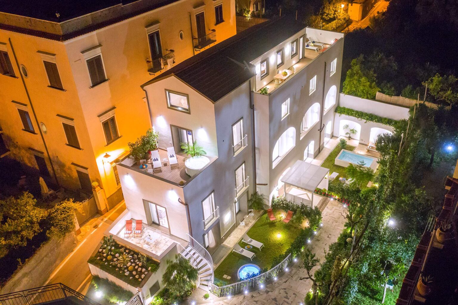 Aerial view of a modern luxury villa illuminated at night, featuring a spacious garden, swimming pool, and multiple outdoor seating areas, surrounded by lush greenery. | Only on SopranoVillas