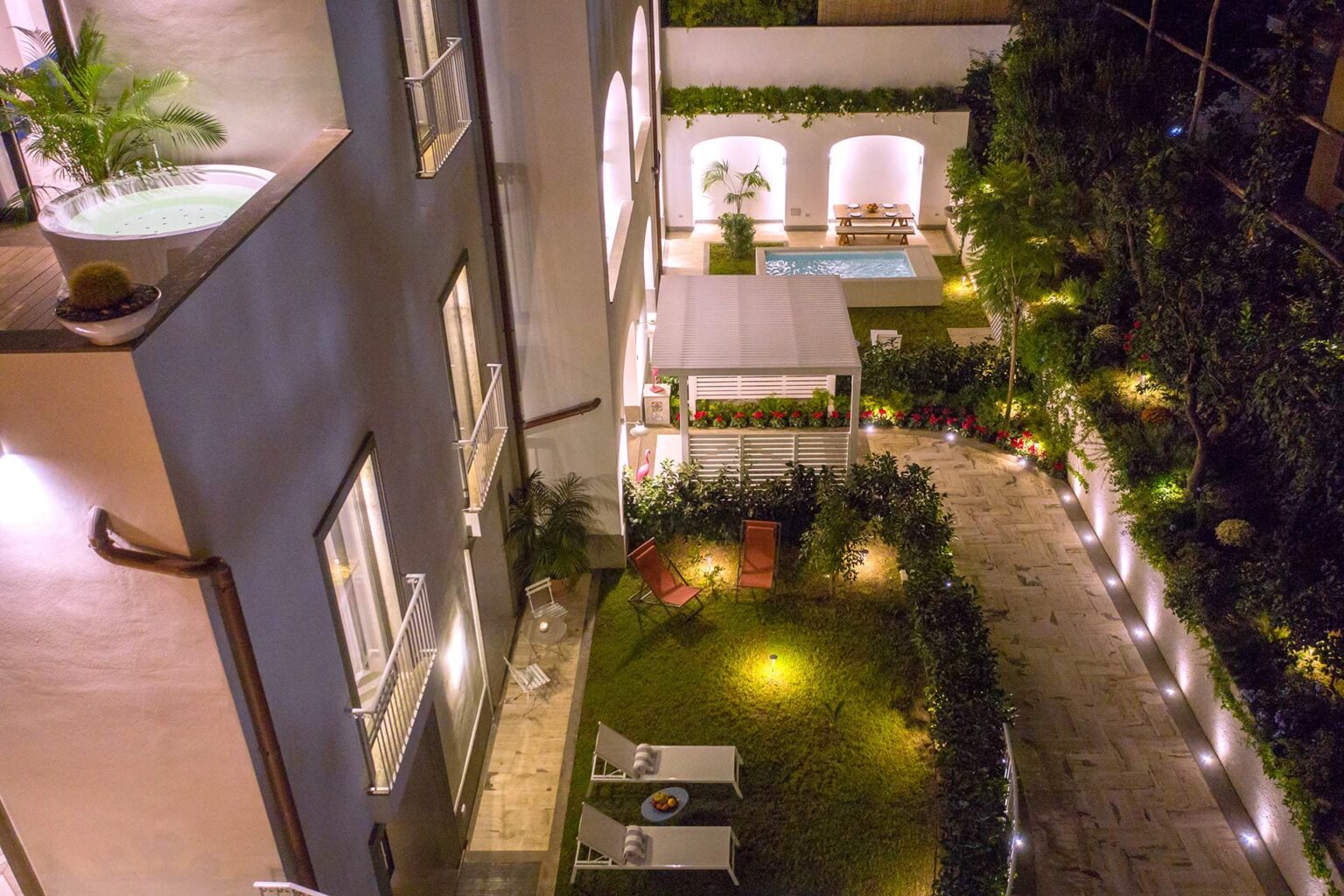 Aerial view of a beautifully landscaped courtyard featuring a swimming pool, lounge chairs, and ambient lighting, surrounded by lush greenery and modern architecture. | Only on SopranoVillas