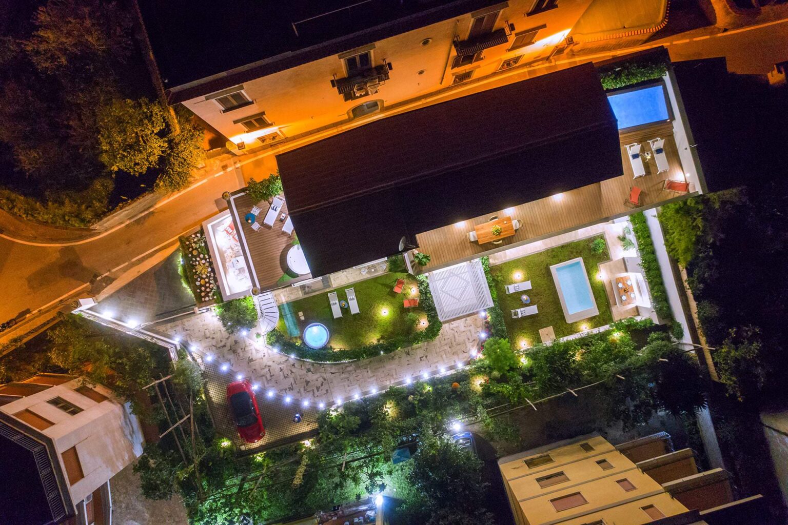 Aerial view of a beautifully lit outdoor space at night, featuring a modern house with a swimming pool, hot tub, and landscaped garden. The area includes comfortable lounge chairs, dining furniture, and pathway lighting, creating a serene ambiance for relaxation and entertainment. | Only on SopranoVillas