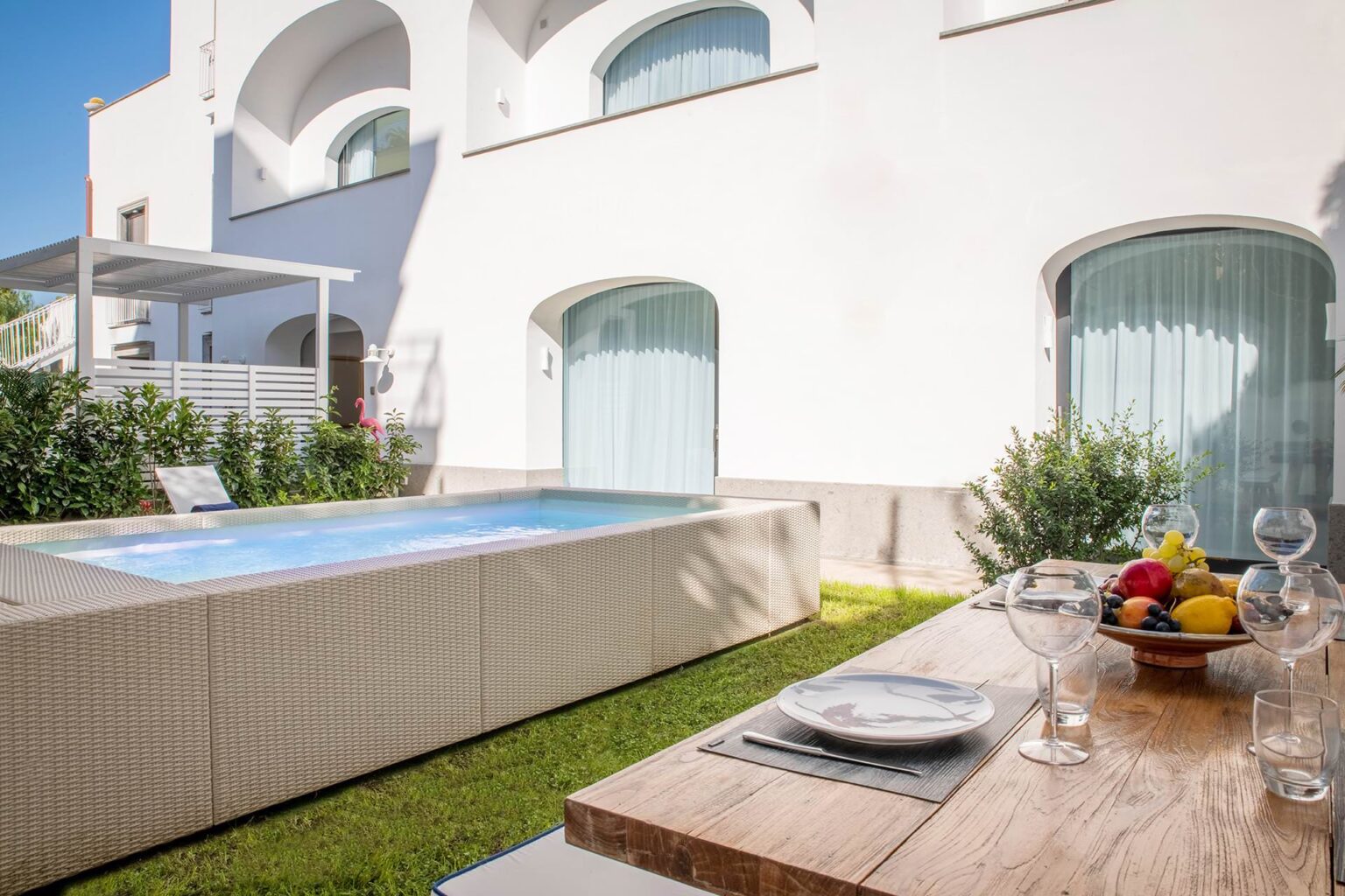 Modern outdoor space featuring a private swimming pool, a wooden dining table set with fruit and glassware, and a backdrop of a stylish white building with large windows. Ideal for relaxation and entertaining in a sunny setting. | Only on SopranoVillas