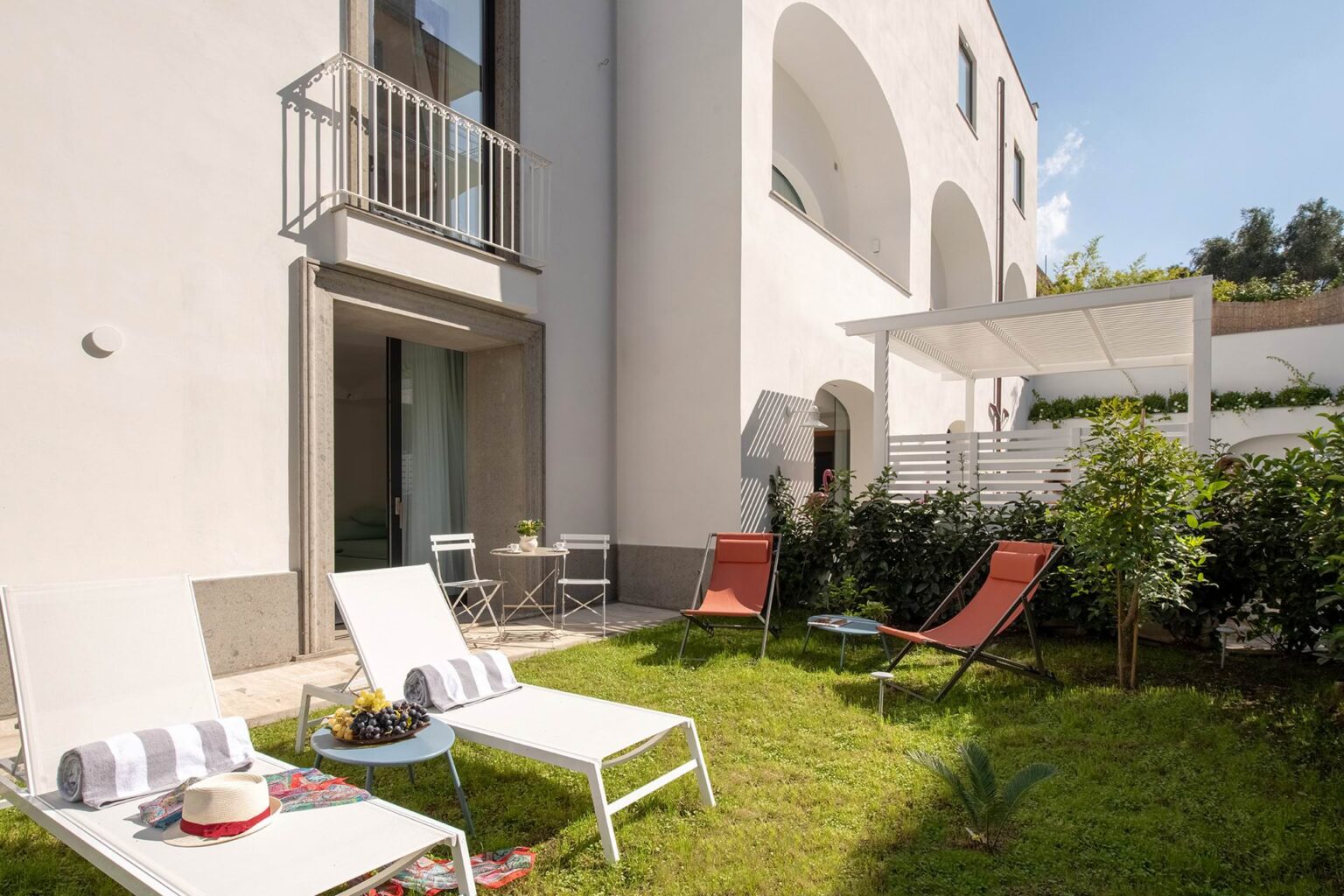 Modern outdoor patio area featuring lounge chairs, a small table, and lush greenery in a sunny setting. The elegant white building has a balcony and large windows, creating a stylish and inviting atmosphere for relaxation. | Only on SopranoVillas