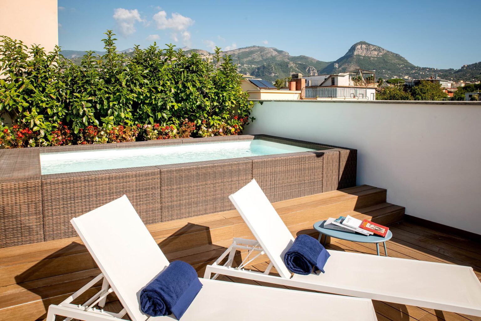 Rooftop terrace featuring a small jacuzzi, two white lounge chairs with blue towels, and a table with books, surrounded by lush greenery and mountains in the background. Ideal setting for relaxation and leisure. | Only on SopranoVillas
