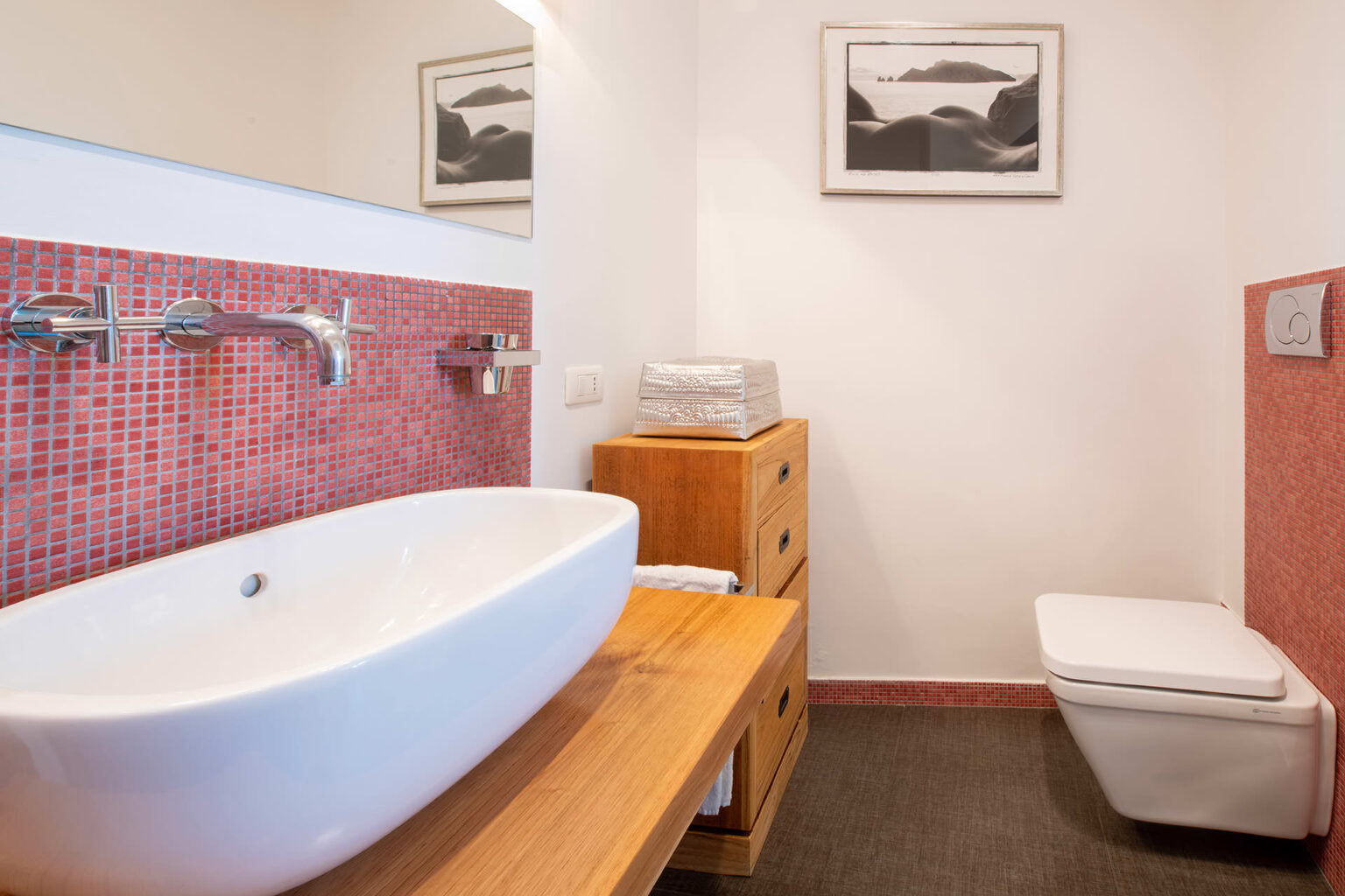 Modern bathroom featuring a freestanding white bathtub, sleek chrome faucets, and a vibrant red mosaic accent wall. The space includes a wooden vanity with storage drawers and a stylish silver decorative box, complemented by a minimalist toilet and a framed artwork on the wall. | Only on SopranoVillas