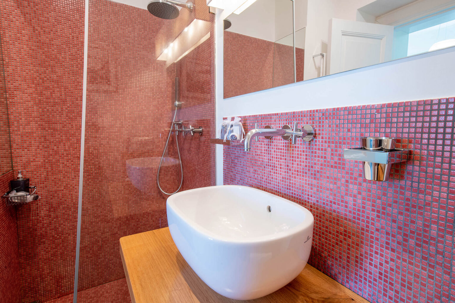 Modern bathroom design featuring a stylish white vessel sink on a wooden countertop, complemented by a vibrant red mosaic tiled wall. The shower area includes a sleek rain showerhead and contemporary fixtures, creating a luxurious and inviting atmosphere. | Only on SopranoVillas