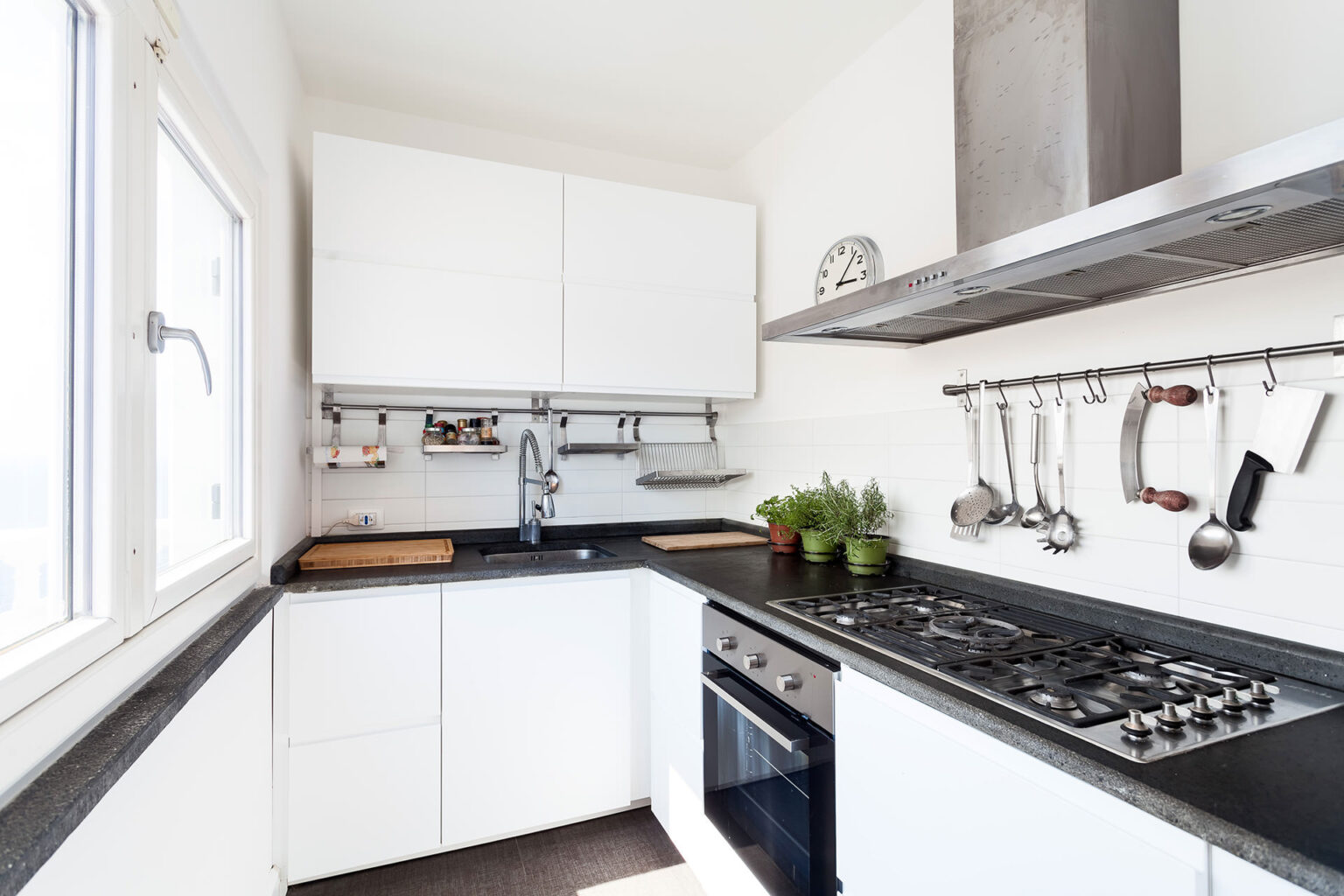 Modern kitchen design featuring sleek white cabinets, a black countertop, stainless steel appliances, and a variety of cooking utensils. The image showcases an organized space with a window providing natural light and potted herbs adding a touch of greenery. | Only on SopranoVillas