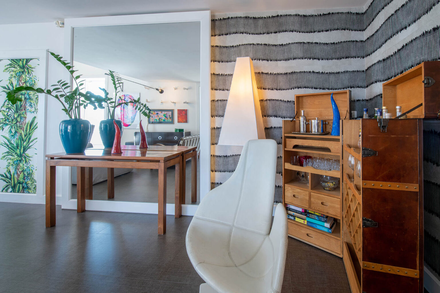Modern living room interior featuring a wooden dining table with potted plants, a stylish white chair, a unique wall lamp, and a wooden bar cabinet displaying glassware and books, complemented by contemporary artwork on the walls. | Only on SopranoVillas