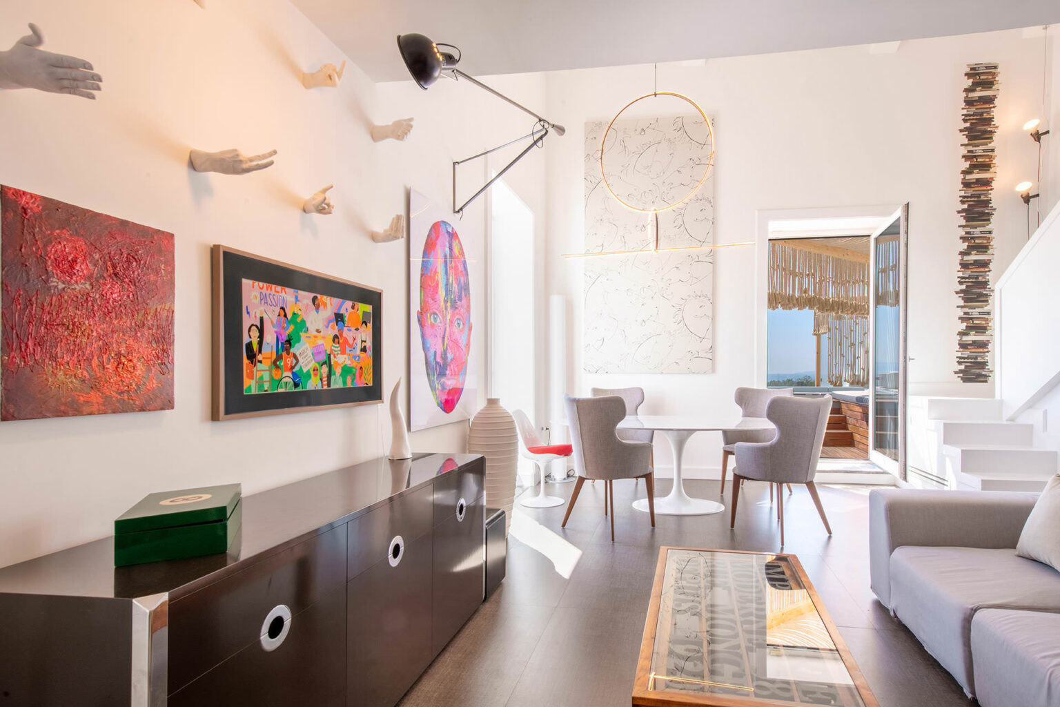 Modern living room featuring eclectic artwork, including colorful paintings and sculptural hands on the wall. The space includes a sleek black cabinet, stylish gray chairs, and a round dining table, with large windows offering a view. Unique lighting fixtures enhance the contemporary design. | Only on SopranoVillas