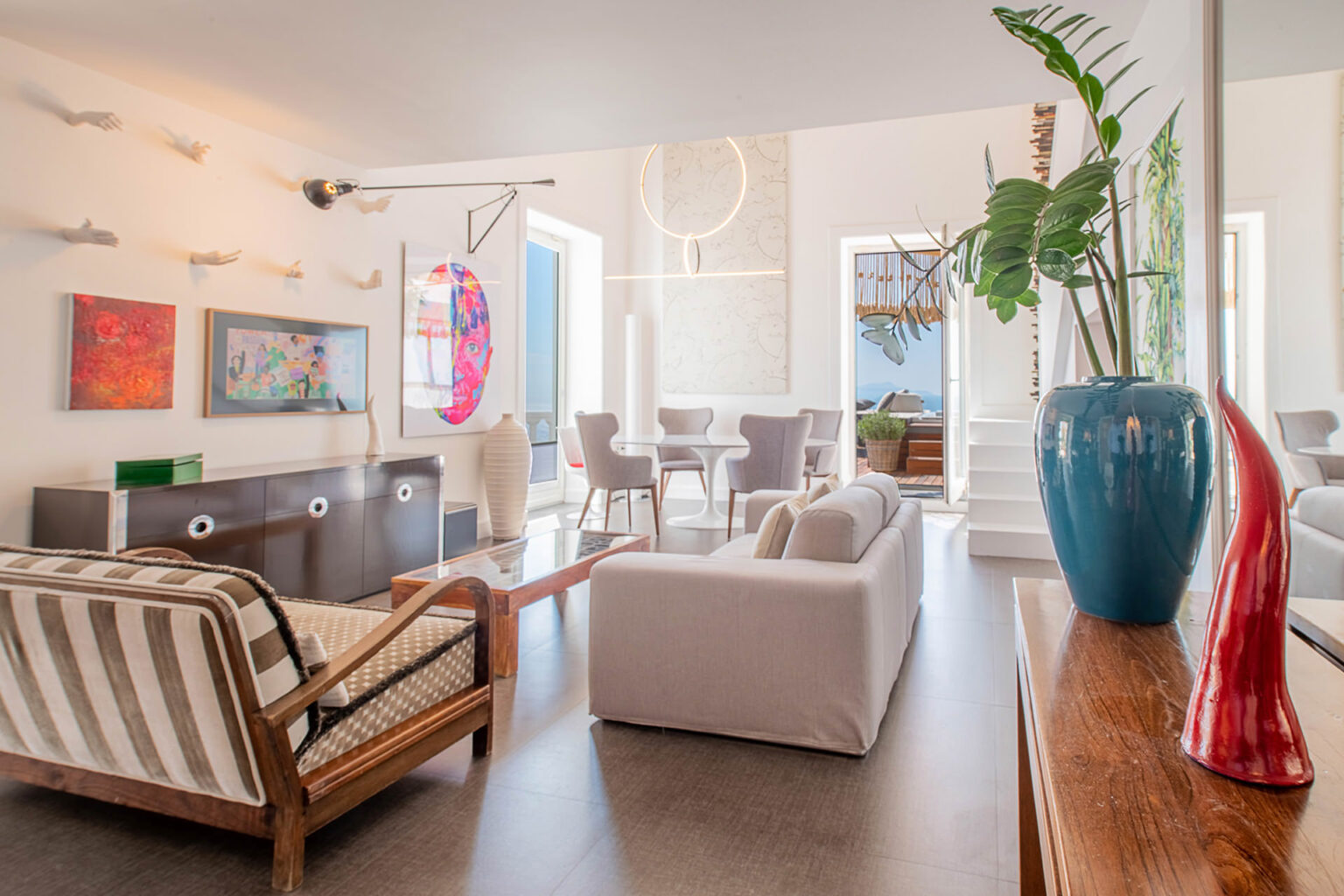 Modern living room featuring stylish furniture, colorful artwork, and unique decor elements. The space includes a comfortable sofa, armchairs, and a wooden coffee table, with large windows allowing natural light. Green plants and decorative vases enhance the contemporary aesthetic. | Only on SopranoVillas
