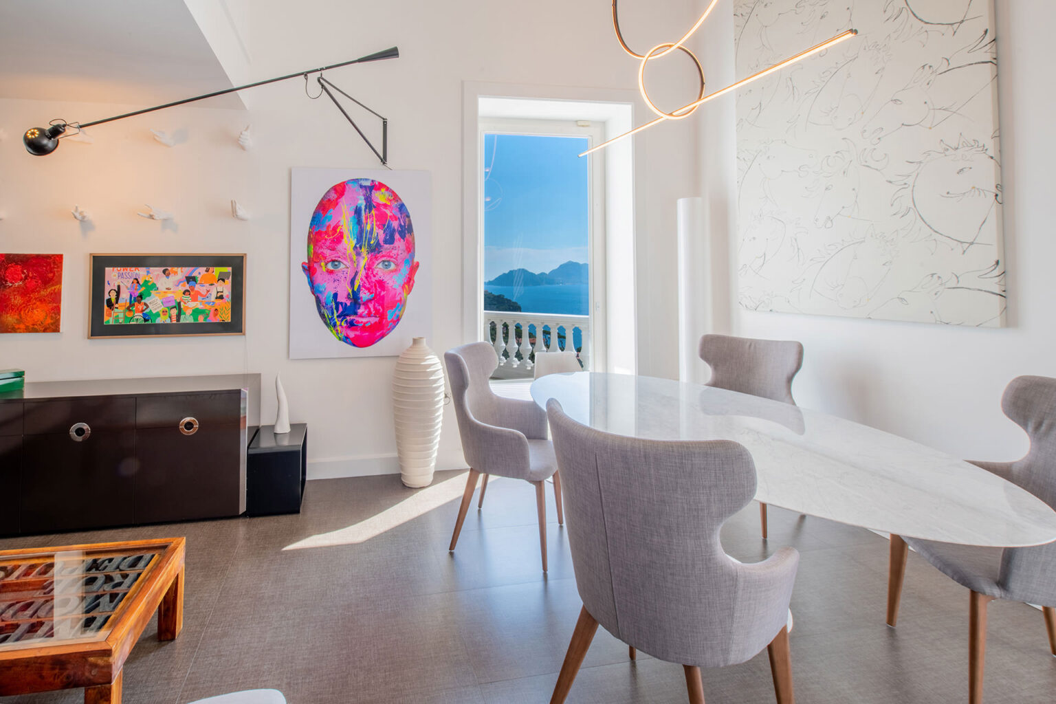 Modern dining room featuring a sleek white table surrounded by stylish gray chairs. Colorful artwork adorns the walls, including a vibrant portrait and abstract pieces. A large window offers a stunning view of mountains and water, while contemporary lighting fixtures add elegance to the space. | Only on SopranoVillas