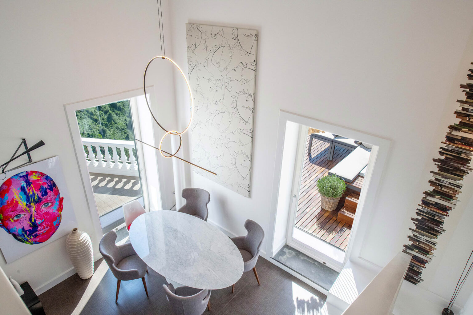 Modern dining area featuring a marble oval table surrounded by gray chairs, illuminated by a contemporary pendant light. A vibrant artwork adorns the wall, and large windows offer a view of the outdoor terrace with wooden flooring and potted greenery. A unique bookshelf design runs vertically along one wall, enhancing the stylish interior. | Only on SopranoVillas