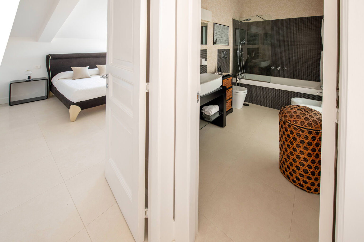 Modern bedroom with a view into an adjoining bathroom, featuring a sleek design, neutral tones, and stylish decor elements like a patterned ottoman and contemporary fixtures. | Only on SopranoVillas
