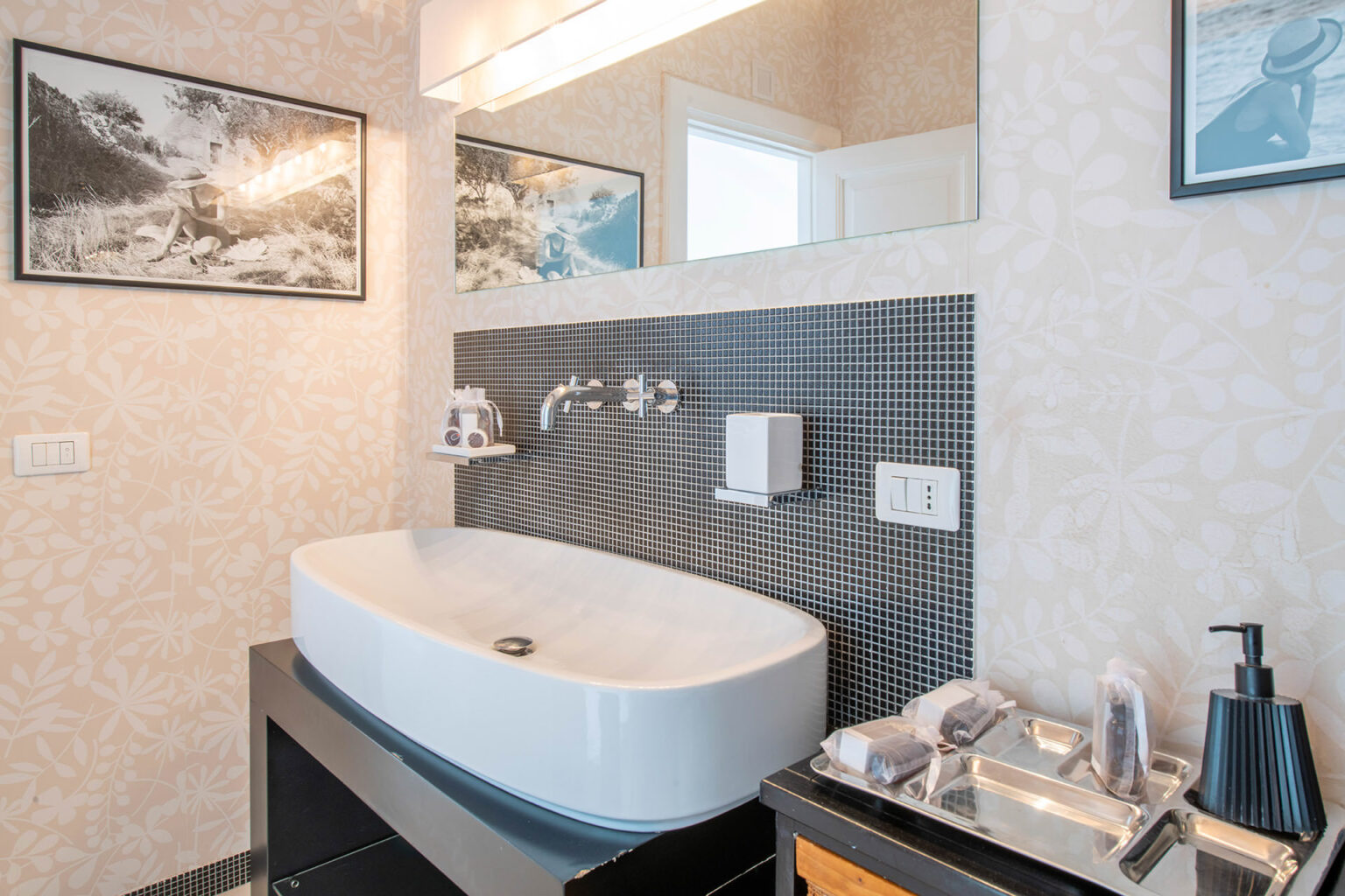 Modern bathroom featuring a sleek freestanding bathtub, stylish wall-mounted faucets, and a decorative mirror. The walls are adorned with textured wallpaper and framed art, while the vanity area includes organized toiletries and a soap dispenser. Natural light enhances the contemporary design. | Only on SopranoVillas