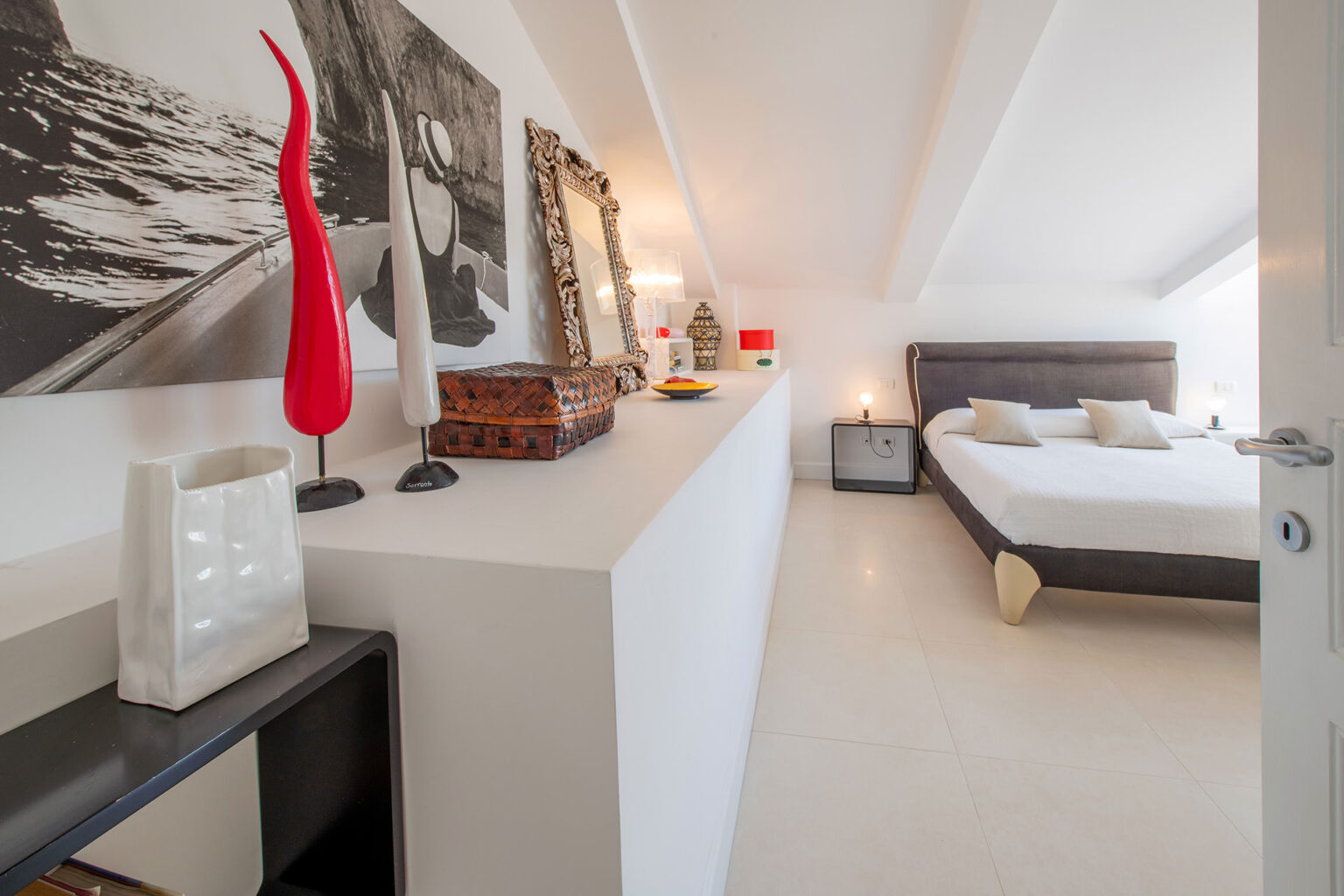 Modern attic bedroom featuring a sleek design with a wooden bed, decorative artwork, and stylish accessories. The room is illuminated by natural light and has a minimalist aesthetic, showcasing a blend of comfort and elegance. | Only on SopranoVillas