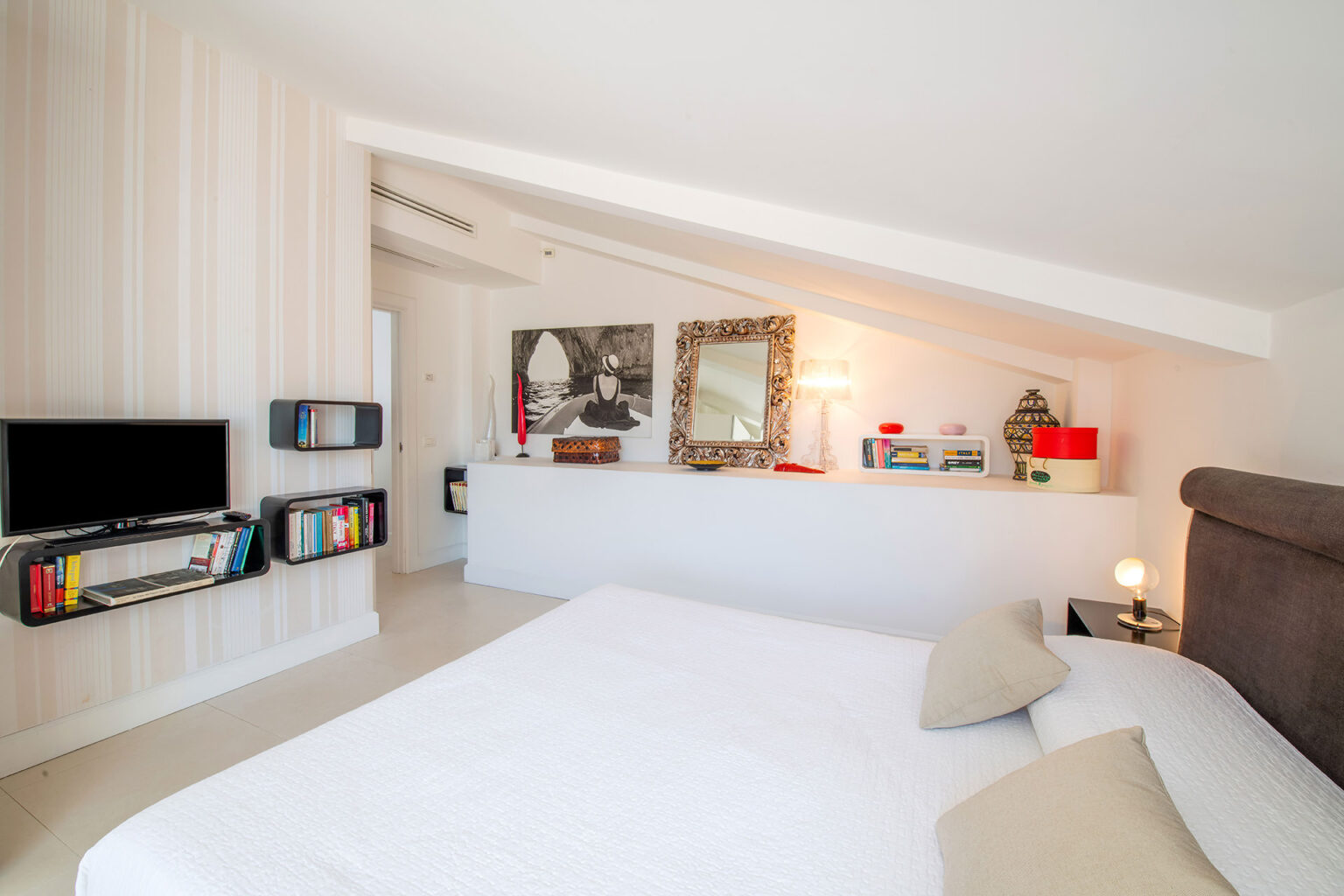 Modern bedroom with sloped ceiling, featuring a comfortable bed, stylish decor, a flat-screen TV, and bookshelves. The space is bright and inviting, with neutral tones and decorative mirrors enhancing the ambiance. | Only on SopranoVillas