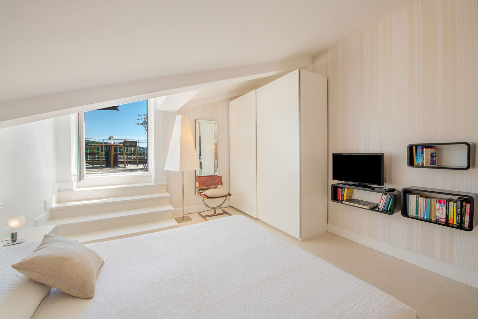 Modern bedroom interior featuring a minimalist design, large windows with a view, a white bed, stylish chair, built-in wardrobe, and wall-mounted shelves with books. The bright, airy space emphasizes natural light and contemporary decor. | Only on SopranoVillas