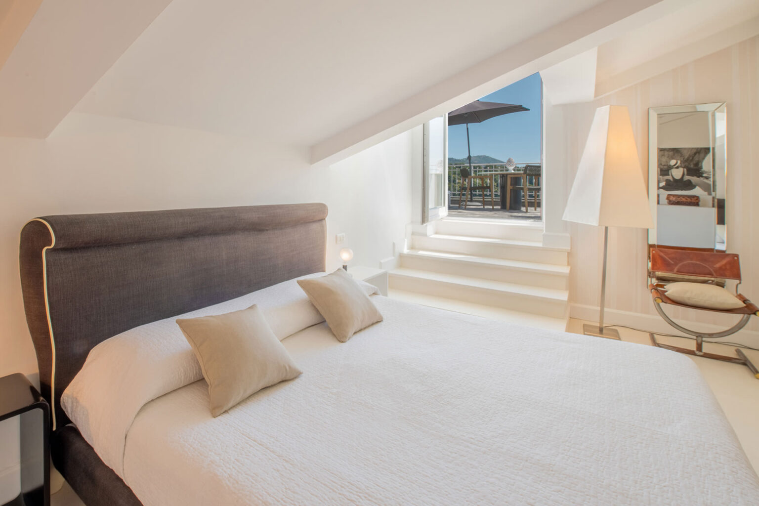 Cozy and modern bedroom featuring a comfortable bed with decorative pillows, large windows providing natural light and city views, and stylish decor including a mirror and a chair, ideal for relaxation and comfort. | Only on SopranoVillas