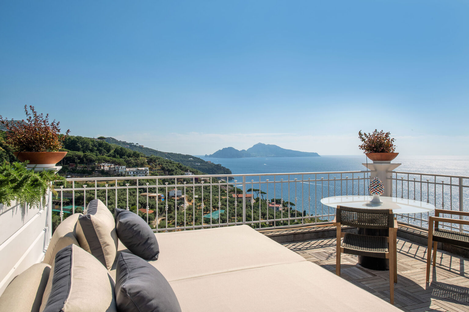 Scenic view from a terrace overlooking the coastline and mountains, featuring a comfortable outdoor seating area with cushions and a round table, perfect for relaxation in a sunny environment. | Only on SopranoVillas