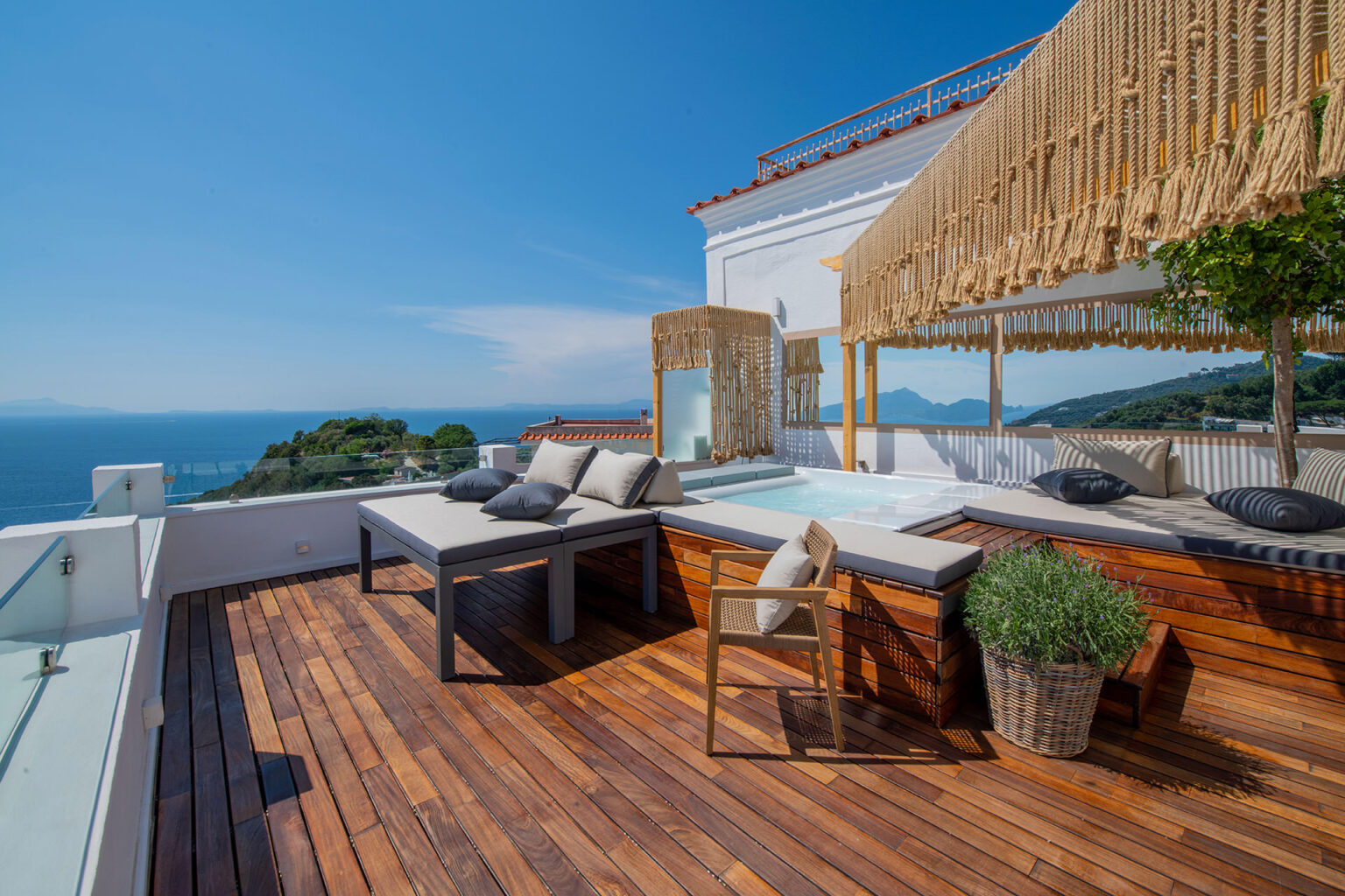 Luxurious rooftop terrace with wooden flooring, comfortable seating, and a hot tub overlooking the ocean, featuring a clear blue sky and lush greenery in the background. Perfect for relaxation and outdoor entertaining. | Only on SopranoVillas