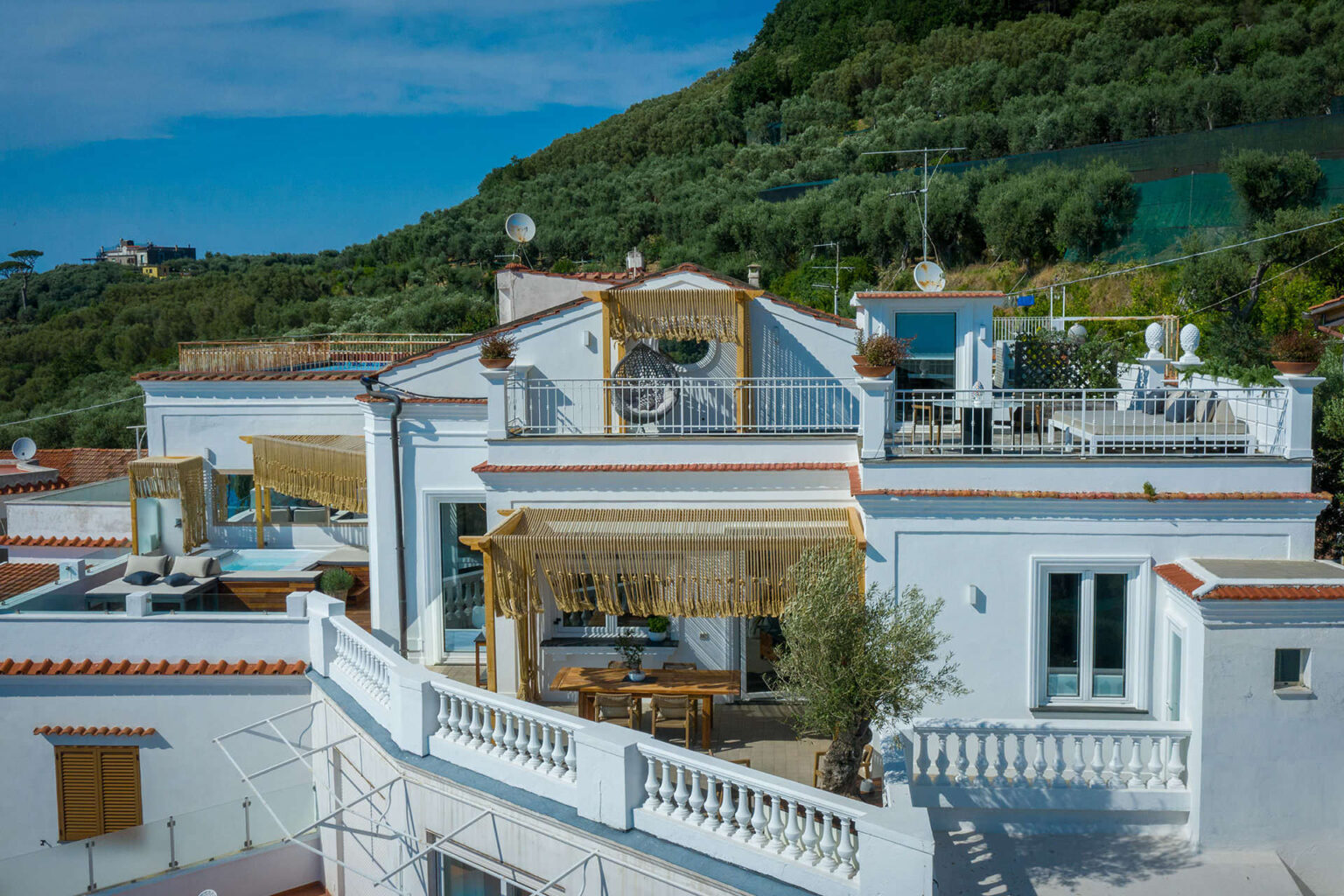 Aerial view of a modern white villa with terracotta roofing, featuring outdoor seating areas, decorative plants, and a lush green hillside in the background. The property showcases stylish design elements, including a covered terrace and balcony space. | Only on SopranoVillas