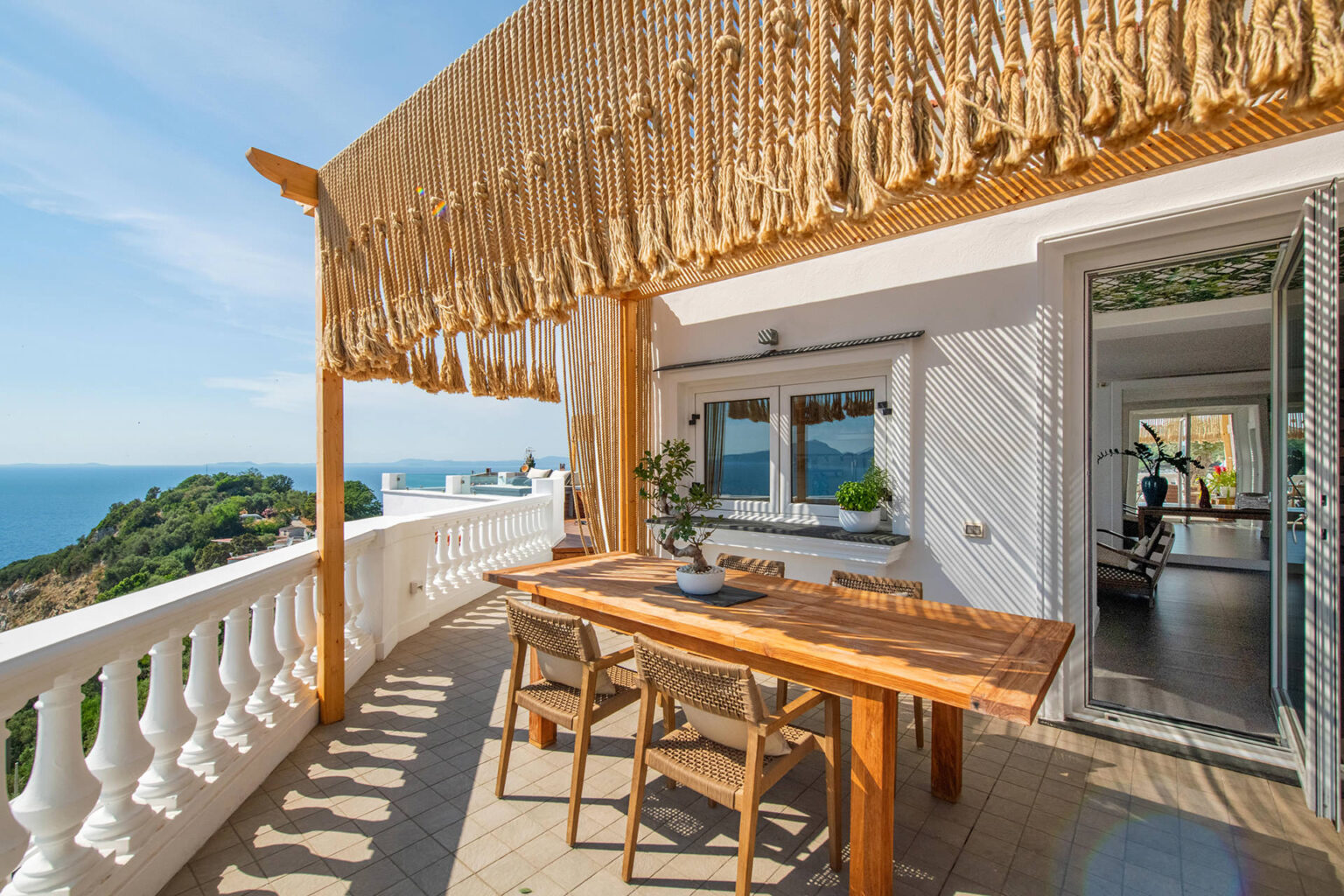 Spacious outdoor terrace with a wooden dining table and chairs, featuring a decorative rope canopy, overlooking a scenic view of the ocean and lush greenery. Ideal for outdoor dining and relaxation. | Only on SopranoVillas