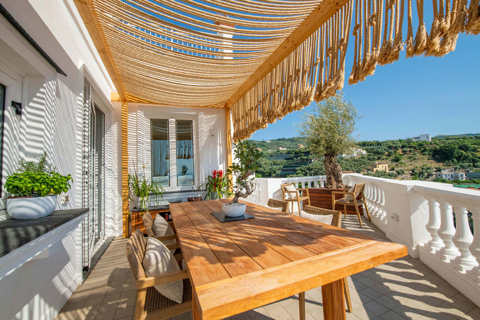 Spacious outdoor terrace featuring a wooden dining table, comfortable seating, and decorative plants, with a stylish rope canopy providing shade and a scenic view of lush green hills and blue sky. | Only on SopranoVillas
