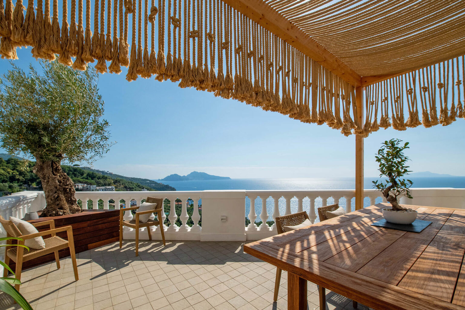 Scenic terrace view overlooking the sea, featuring a wooden dining table, comfortable chairs, and an olive tree, with a decorative canopy providing shade against a clear blue sky. | Only on SopranoVillas