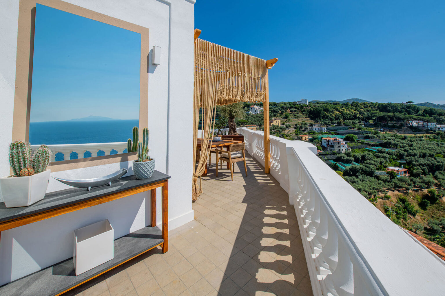 Spacious balcony with stunning sea views, featuring a large mirror, decorative cacti, and a stylish wooden table set under a modern canopy. Ideal for outdoor relaxation and dining in a scenic location. | Only on SopranoVillas
