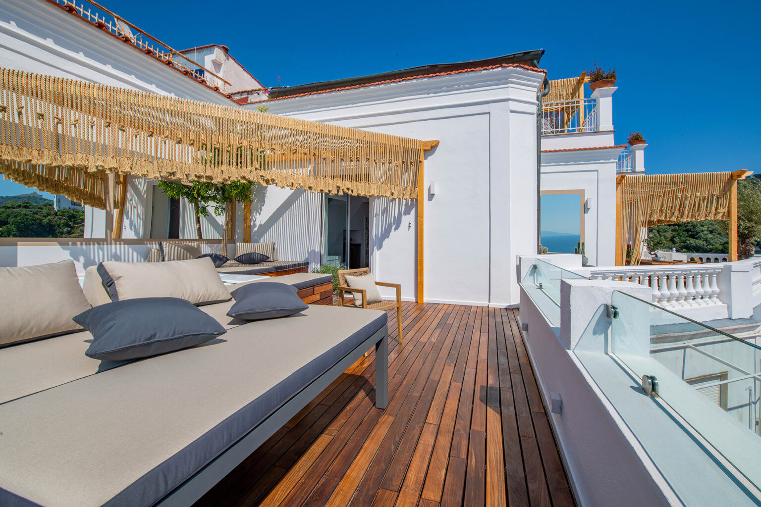 Spacious outdoor terrace featuring a comfortable lounge area with a wooden deck, stylish seating, and a view of the ocean under a clear blue sky. | Only on SopranoVillas