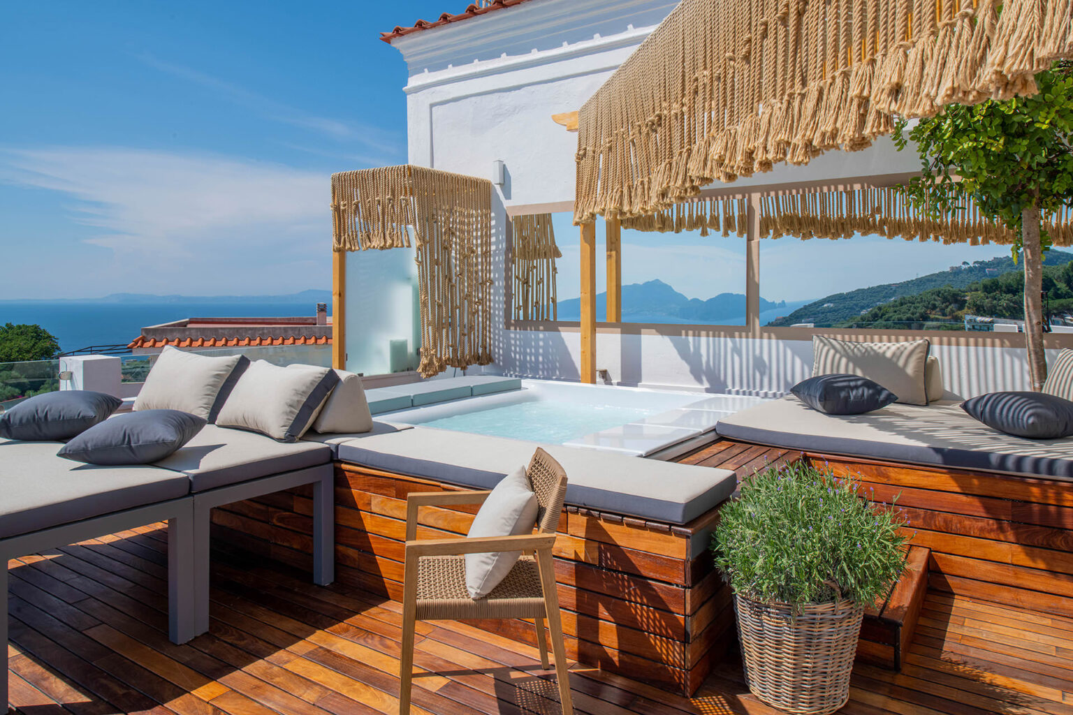 Luxurious outdoor terrace featuring a hot tub, comfortable lounge seating with plush cushions, and a stunning ocean view. The space is adorned with stylish wooden decking and decorative fringed canopies, perfect for relaxation and enjoying scenic landscapes. | Only on SopranoVillas