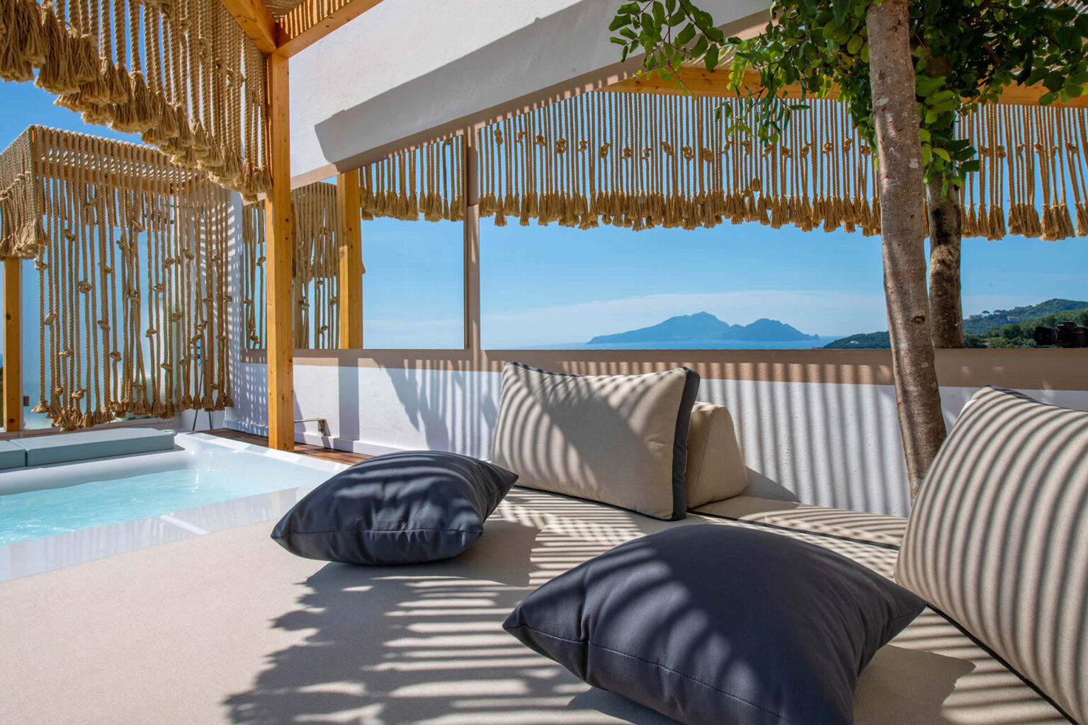 Modern outdoor lounge area with plush cushions, a private plunge pool, and stunning mountain views, featuring stylish wooden decor and vibrant sunlight. | Only on SopranoVillas