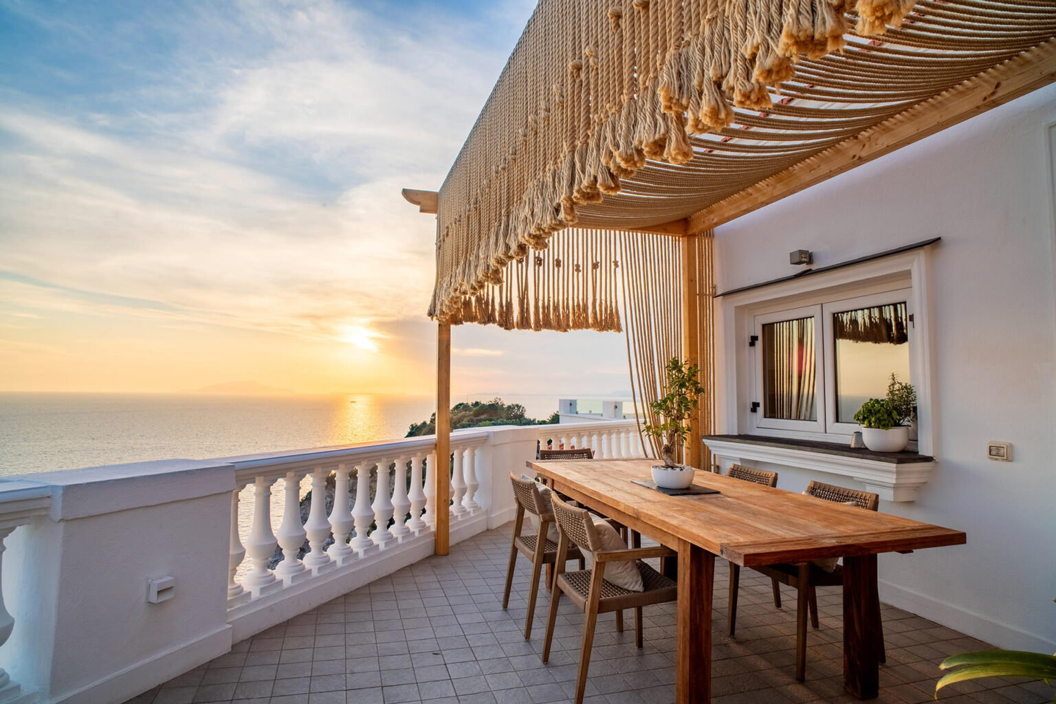 Cozy outdoor dining area on a balcony with a wooden table and chairs, overlooking a sunset over the ocean, featuring a stylish canopy and decorative elements. | Only on SopranoVillas