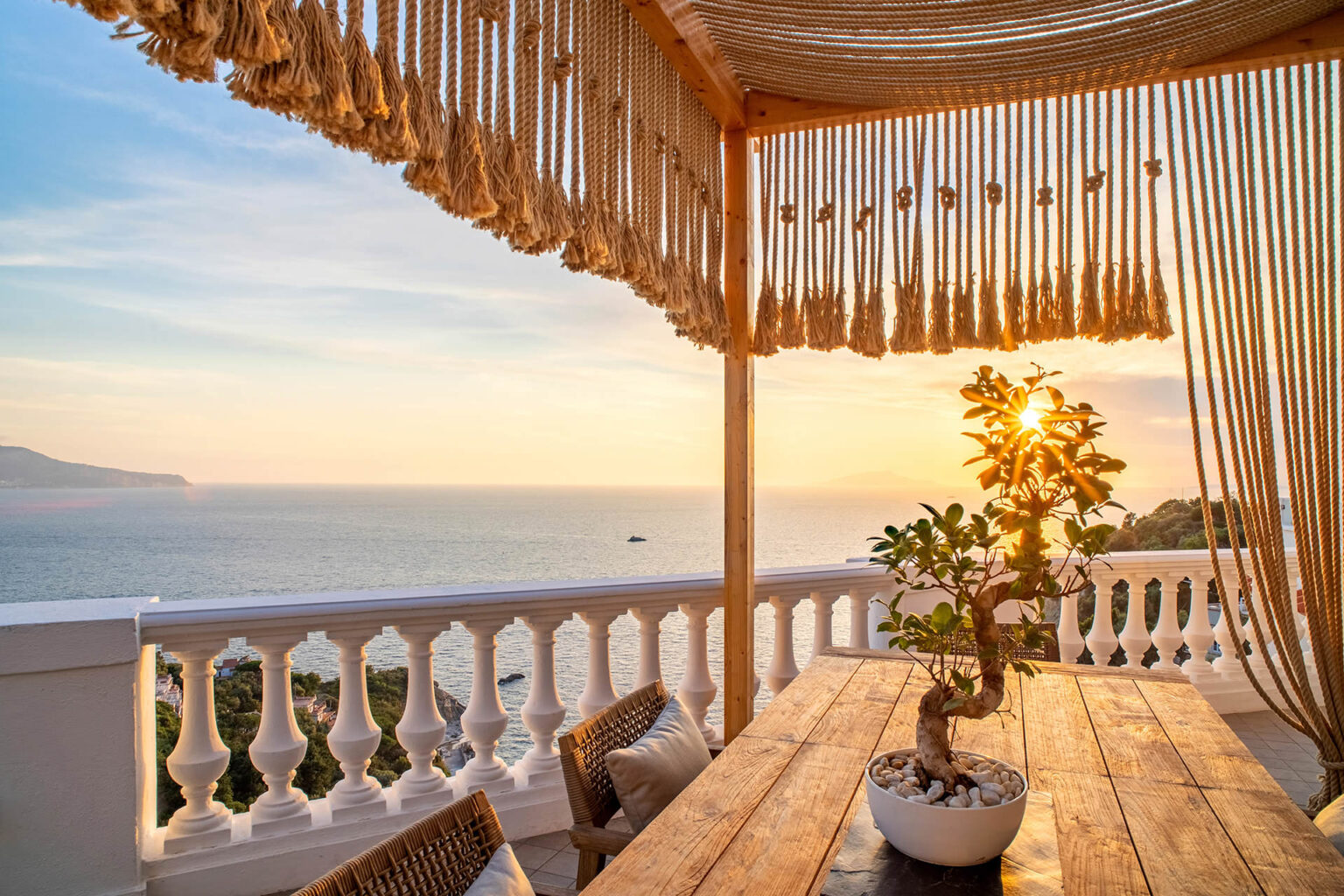 Cozy outdoor terrace with a wooden table and decorative bonsai tree, overlooking a serene ocean sunset, featuring a stylish canopy and comfortable seating. | Only on SopranoVillas
