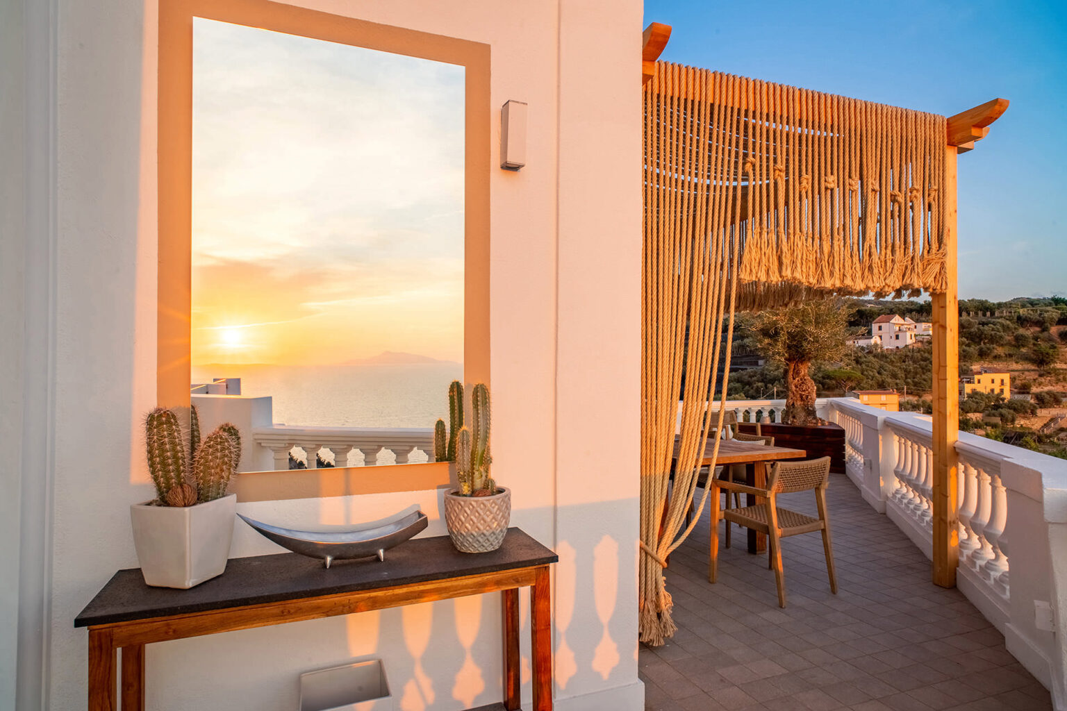 Cozy balcony with a sunset view, featuring a wooden table, potted cacti, and a decorative mirror, perfect for relaxation and outdoor dining. | Only on SopranoVillas