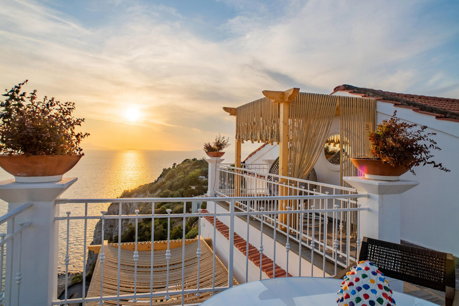 Scenic sunset view from a balcony overlooking the ocean, featuring decorative pots with plants and a cozy seating area, ideal for relaxation and enjoying nature. | Only on SopranoVillas