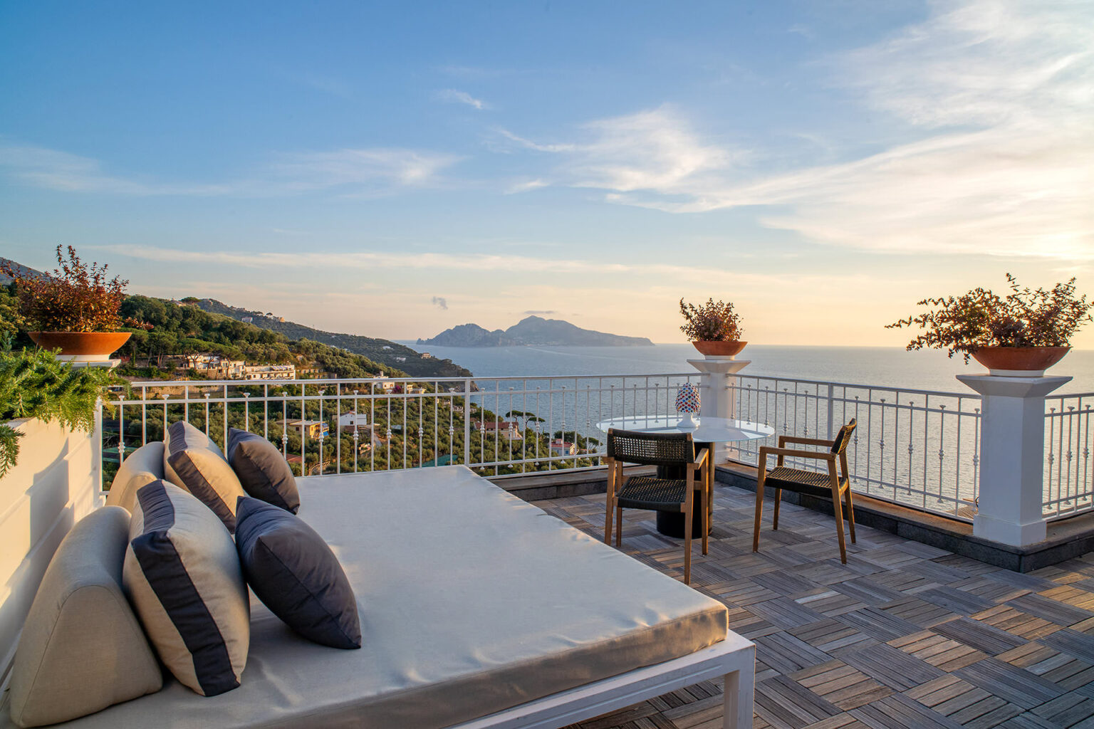 Sunset view from a stylish terrace overlooking the ocean, featuring a comfortable lounge area with cushions and a dining table with chairs, surrounded by lush greenery and decorative plants. | Only on SopranoVillas