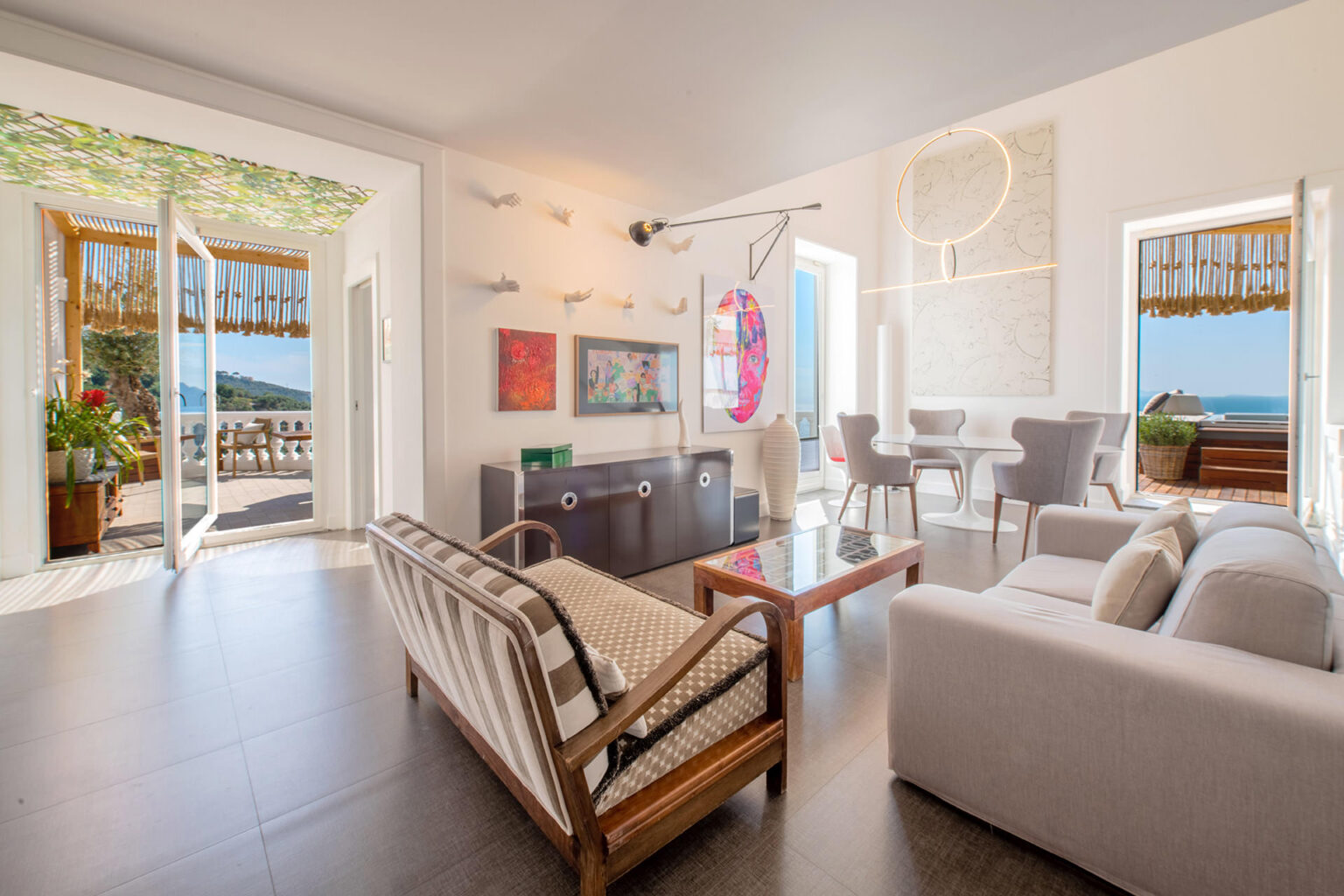 Bright and modern living room with panoramic ocean views, featuring stylish furniture, large windows, and an inviting atmosphere. The space includes a comfortable sofa, wooden chairs, and a dining area, perfect for relaxation and entertaining. | Only on SopranoVillas