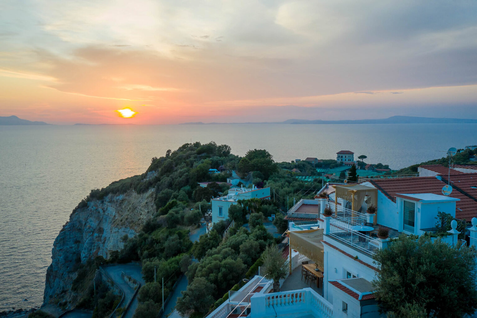 Scenic view of a coastal sunset over the ocean, highlighting cliffs and hillside homes with vibrant colors in the sky. | Only on SopranoVillas