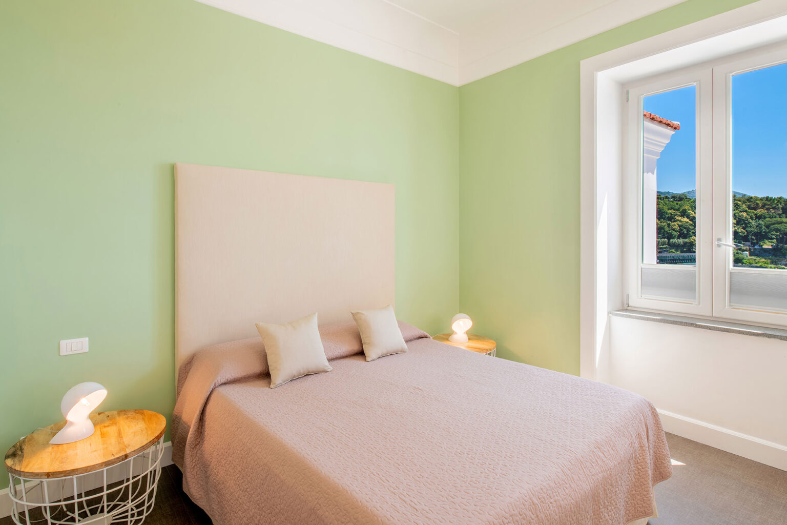 Cozy bedroom featuring a light green accent wall, a queen-sized bed with neutral bedding, modern bedside lamps, and a large window offering scenic views. | Only on SopranoVillas