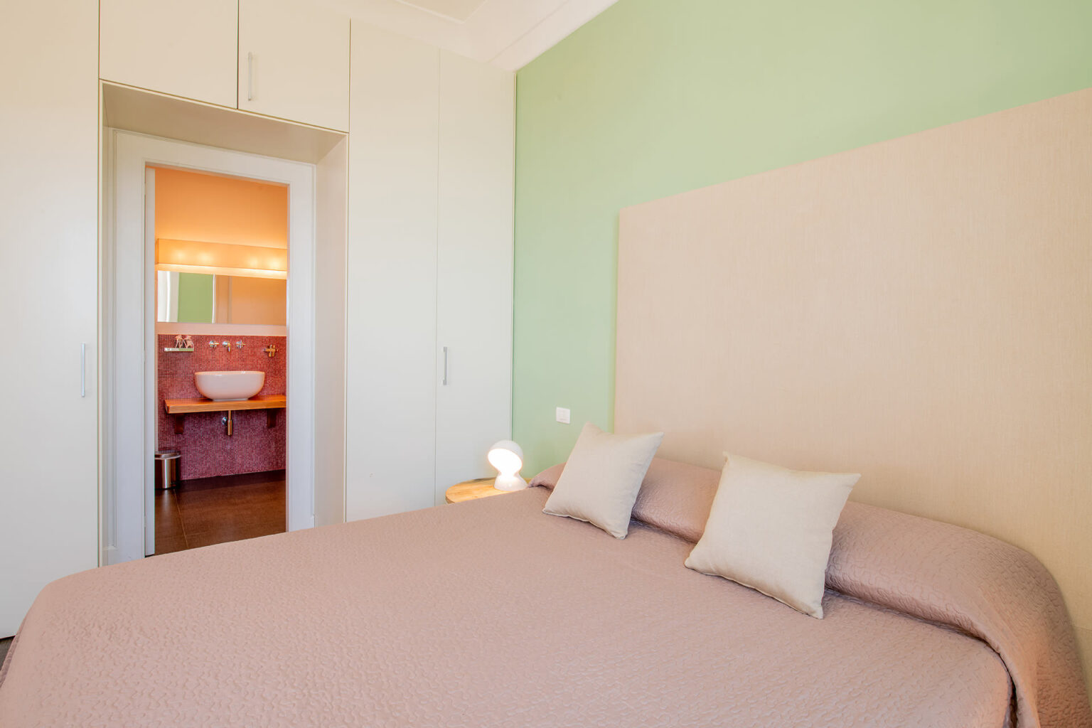 Cozy bedroom featuring a soft pink bedspread, decorative pillows, and a light green accent wall. An open doorway leads to a brightly lit bathroom with a modern sink. Ideal for showcasing interior design and home decor. | Only on SopranoVillas