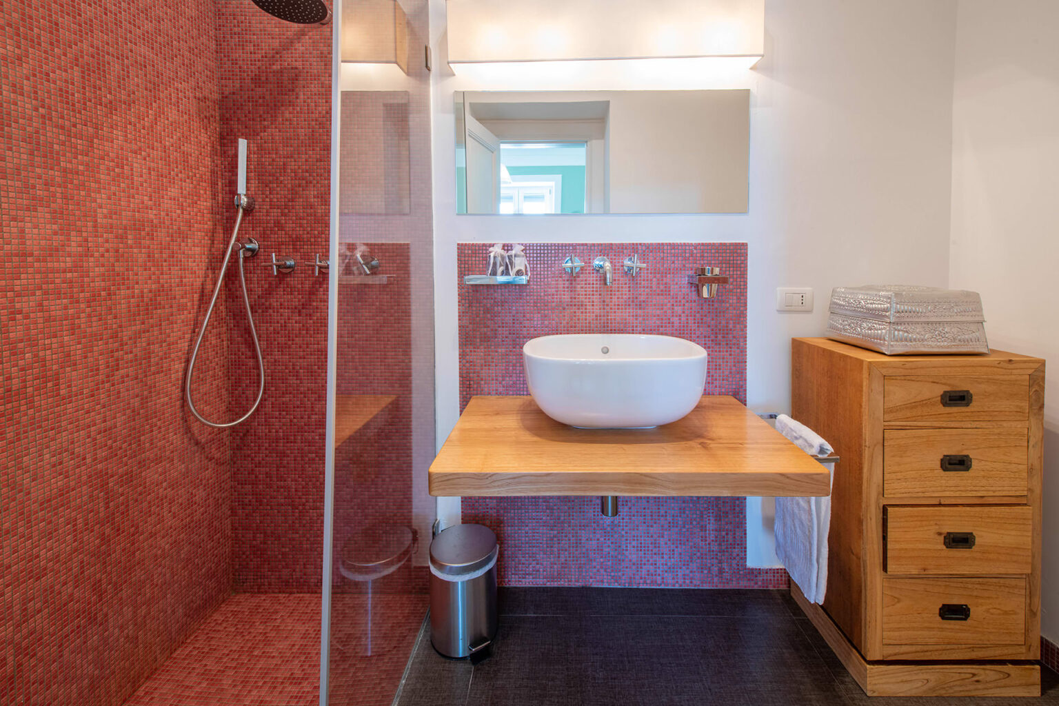 Modern bathroom design featuring a walk-in shower, a stylish vessel sink on a wooden countertop, and wooden storage drawers, all set against a vibrant red wall. | Only on SopranoVillas