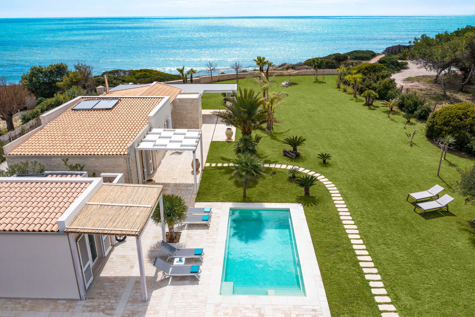 Aerial view of a modern villa with a swimming pool, surrounded by lush green lawn and palm trees, overlooking a serene blue ocean. The image showcases the villa's stylish architecture, outdoor seating areas, and well-maintained landscaping, perfect for relaxation and leisure. | Only on SopranoVillas