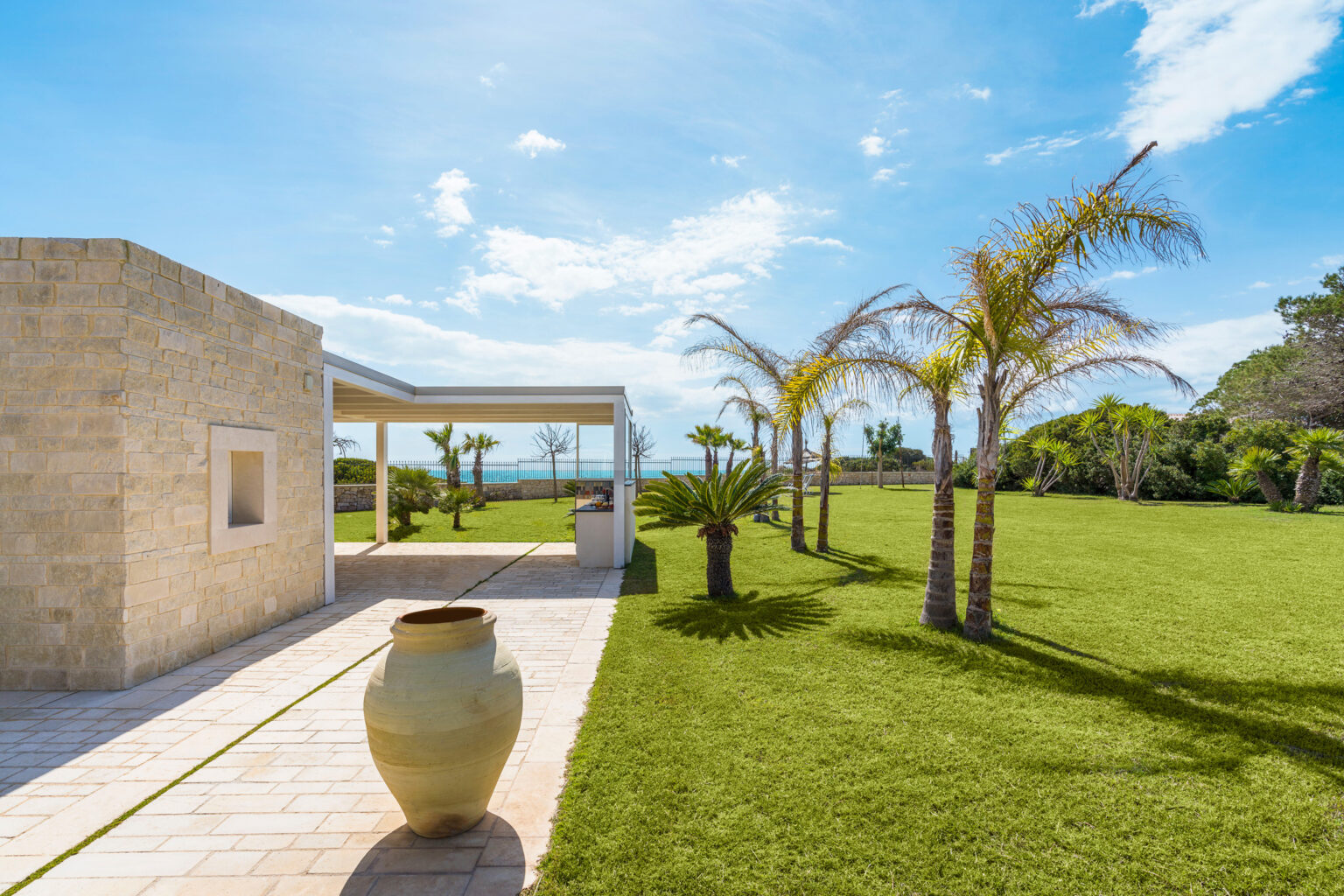 Modern villa exterior with lush green lawn and palm trees under a bright blue sky, featuring a stone pathway and a decorative pot in the foreground. Ideal for luxury real estate listings or vacation property promotions. | Only on SopranoVillas