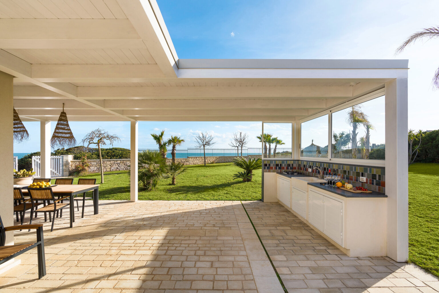 Modern outdoor kitchen and dining area with a view of the ocean, featuring a spacious patio, greenery, and stylish furniture under a covered pergola. | Only on SopranoVillas