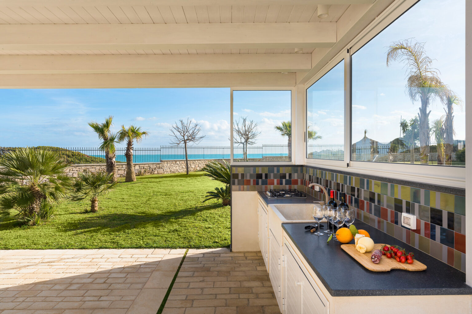 Modern kitchen with colorful backsplash overlooking a serene garden and ocean view, featuring fresh fruits on the countertop. | Only on SopranoVillas