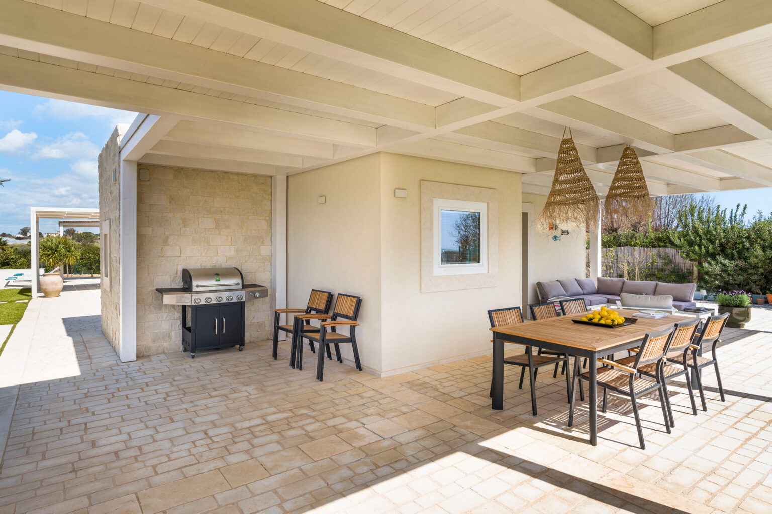 Modern outdoor patio featuring a dining table with chairs, a cozy seating area, and a barbecue grill, surrounded by greenery and under a shaded roof. Ideal for entertaining and enjoying outdoor meals. | Only on SopranoVillas