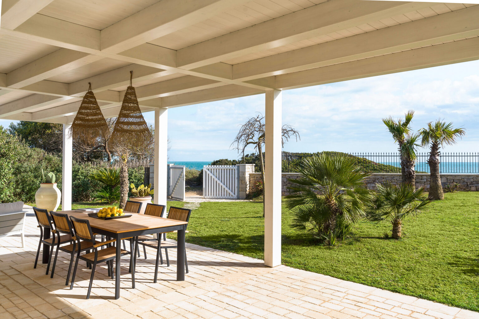 Outdoor dining area with a wooden table and chairs, surrounded by lush greenery and palm trees, overlooking the ocean. The space features a covered patio with hanging light fixtures, creating a serene and inviting atmosphere for al fresco meals. | Only on SopranoVillas
