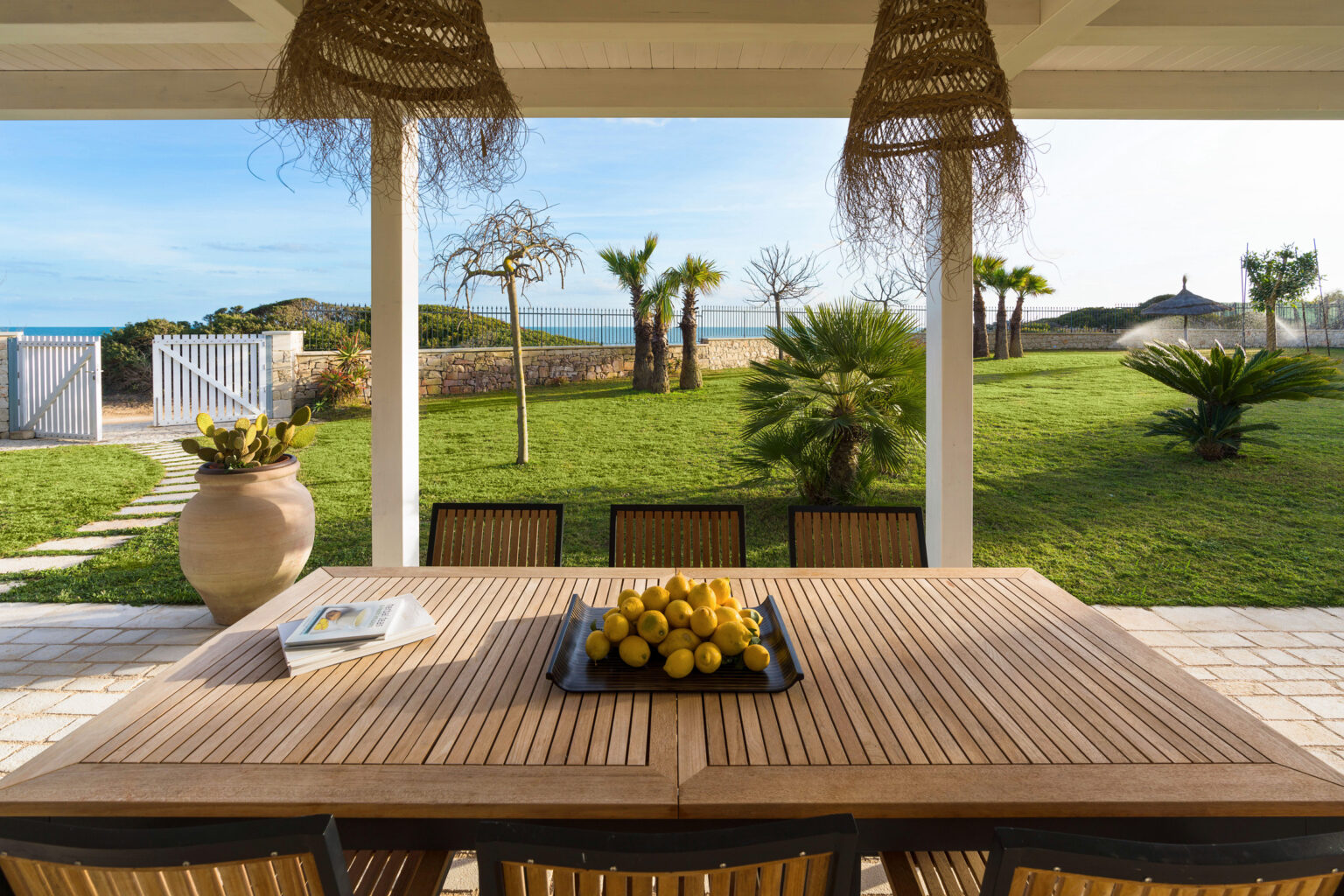 Spacious outdoor dining area with a wooden table adorned with a centerpiece of lemons, surrounded by lush greenery and palm trees, overlooking a serene ocean view. | Only on SopranoVillas