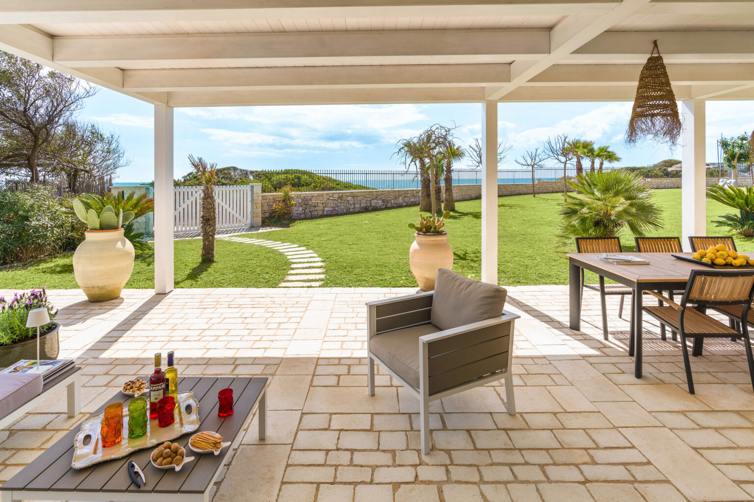 A stylish outdoor patio with comfortable seating, a dining table, and a scenic view of a lush green garden and ocean in the background. The setting features decorative vases, a coffee table with snacks and drinks, and a bright, inviting atmosphere perfect for relaxation and entertaining. | Only on SopranoVillas